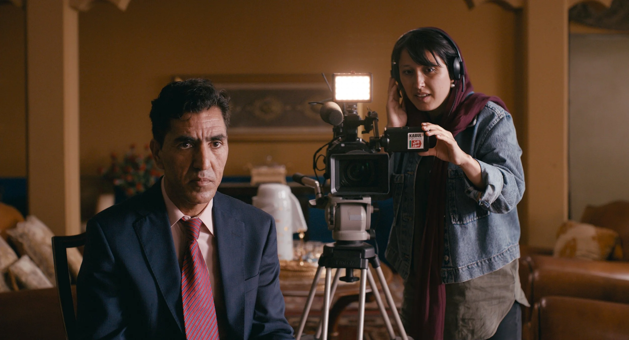 Bilde av en mann som sitter på en stol iført dress. Det er dempet lys. Bak han er en dame som filmer han med et stort filmkamera 