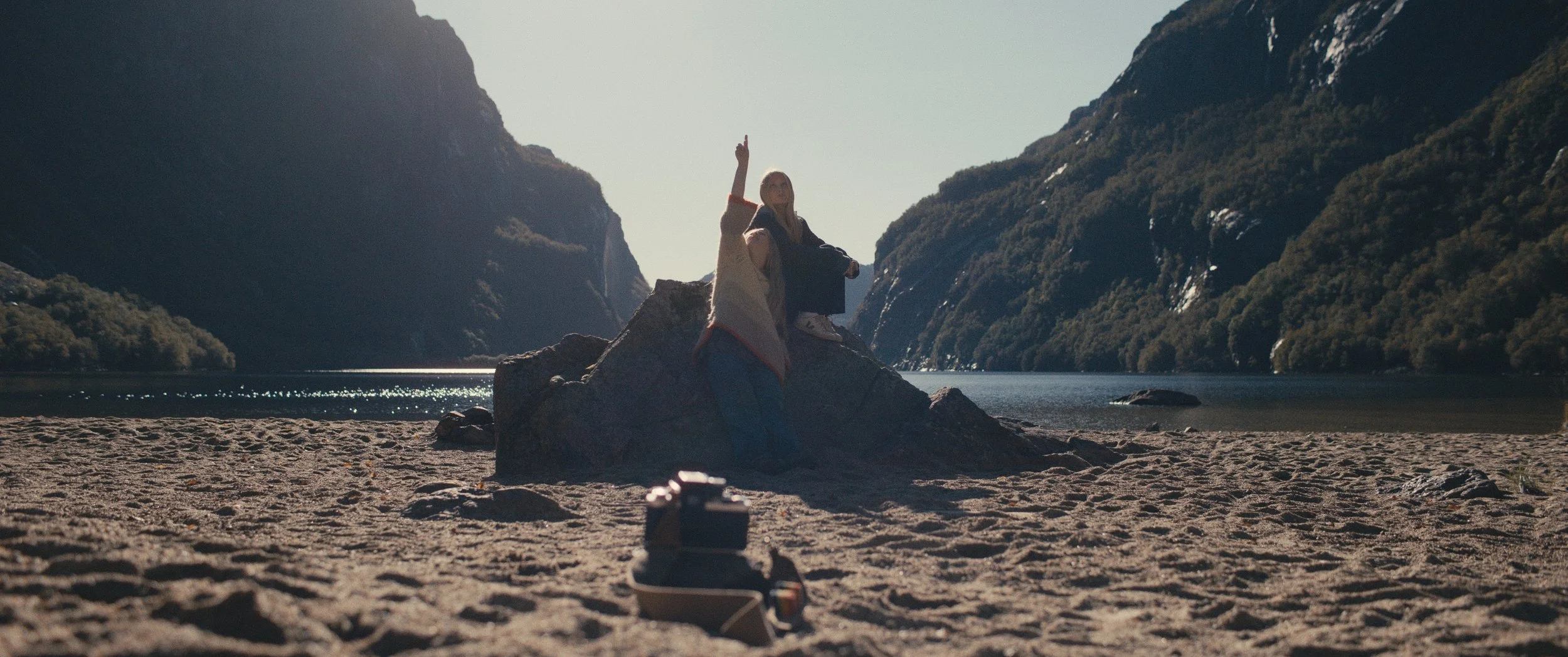 Et landskapsbilde som viser en sandstrand med fjordlandskap rundt seg. Midt i bildet er det en stein der en jente lener seg og peker opp i været. På samme steinen sitter en annen jente som der opp i været der den andre jenten peker.