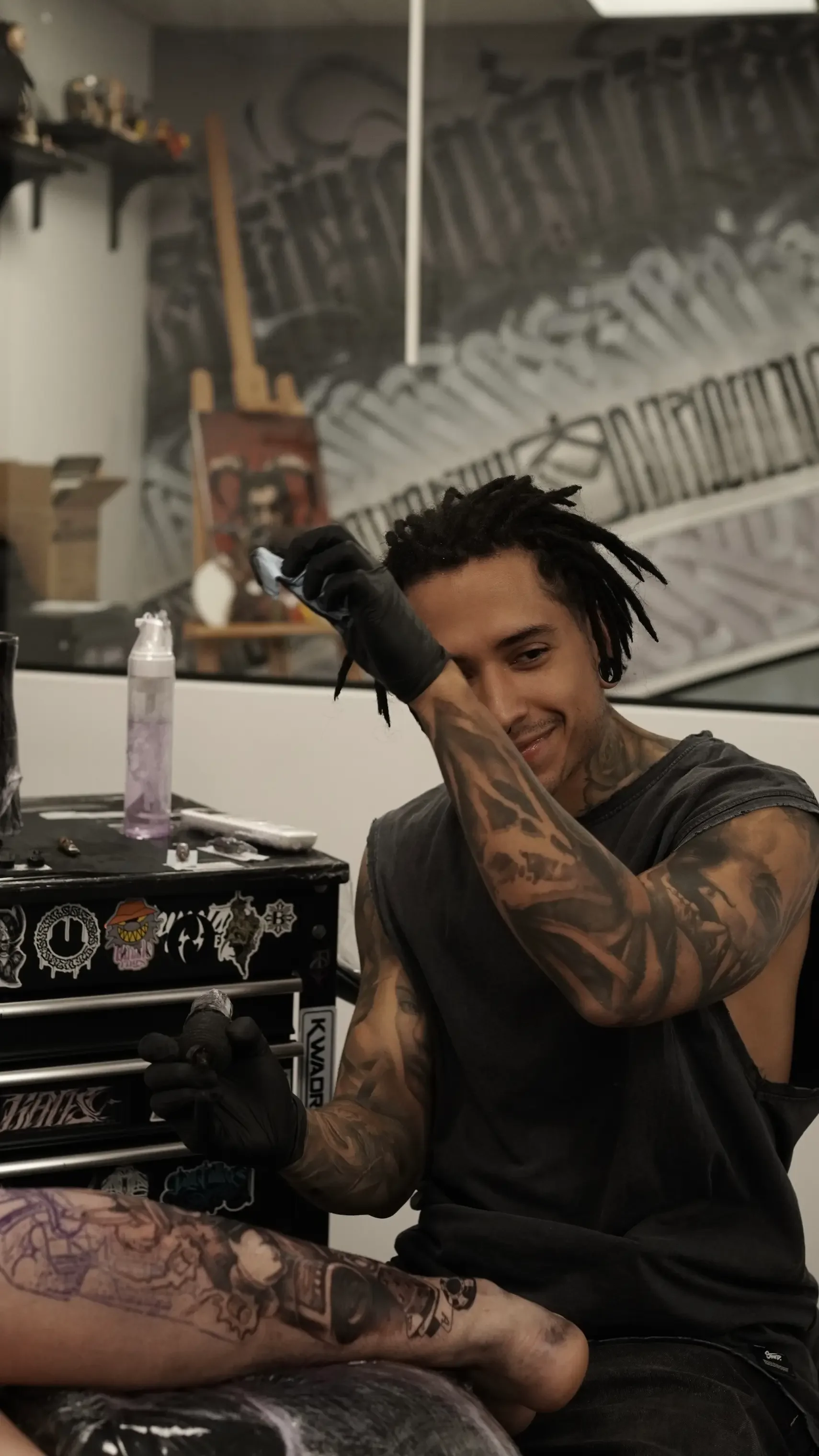 Un hombre con tatuajes y cabello rasta sonríe y se limpia la frente con la mano, sentado en un taller de tatuajes con un área de trabajo y arte en la pared de fondo.