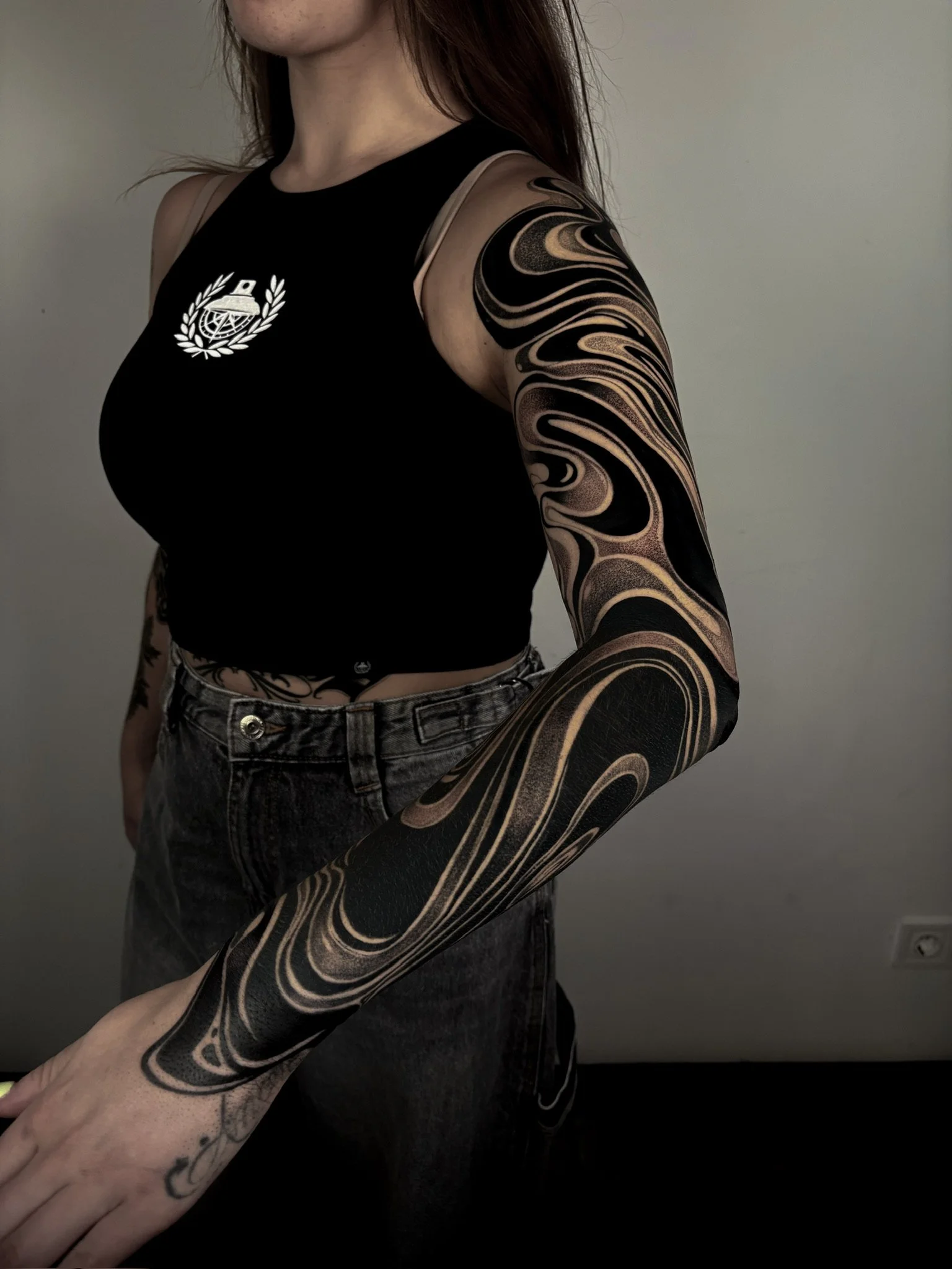 Persona con tatuajes de patrones abstractos en el brazo, usando camiseta negra con logotipo en el pecho y pantalones negros, en un fondo gris.