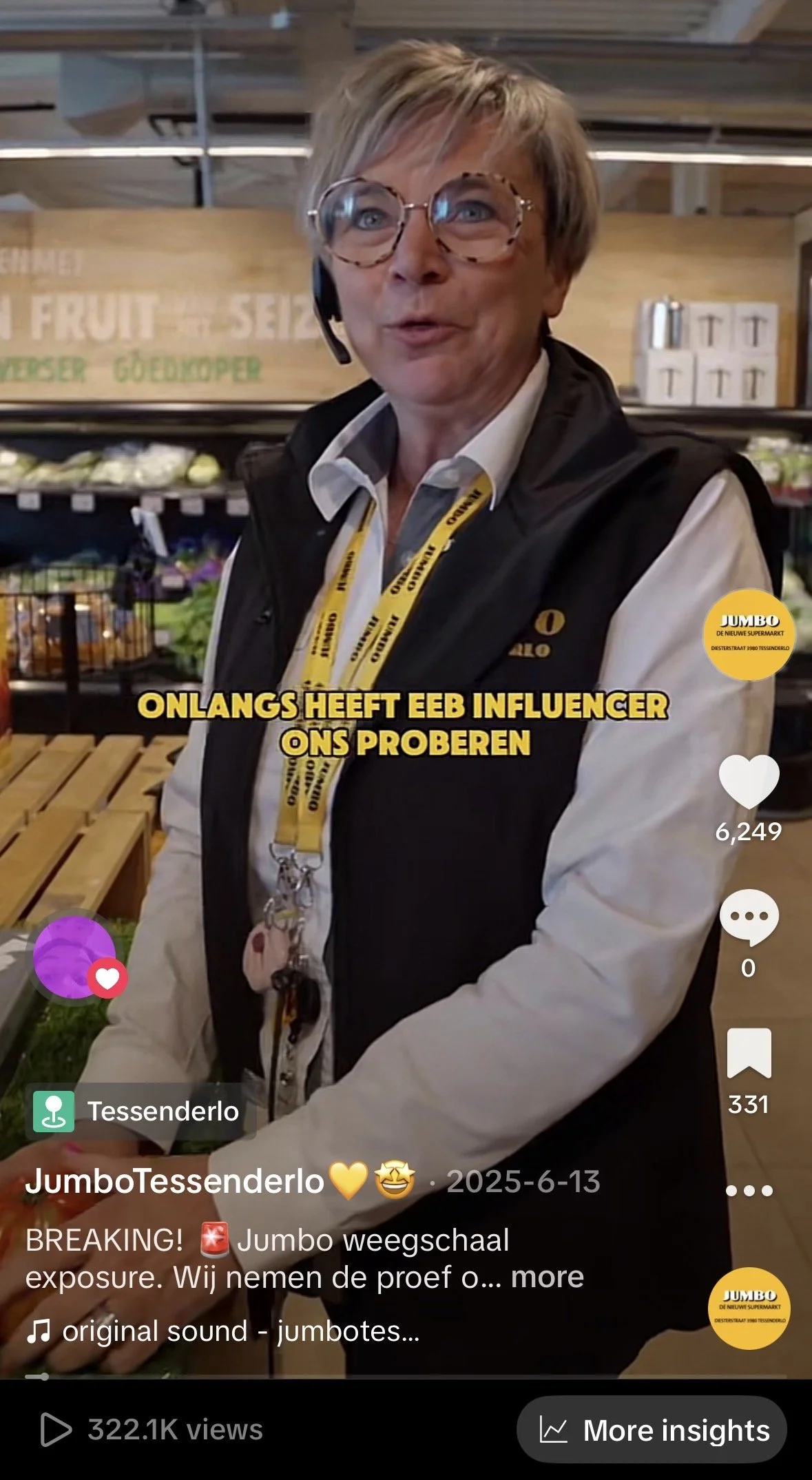 Vrouw met korte haar, bril, headset en Jumbo badge, staand in een supermarkt met groenten en fruit op de achtergrond.