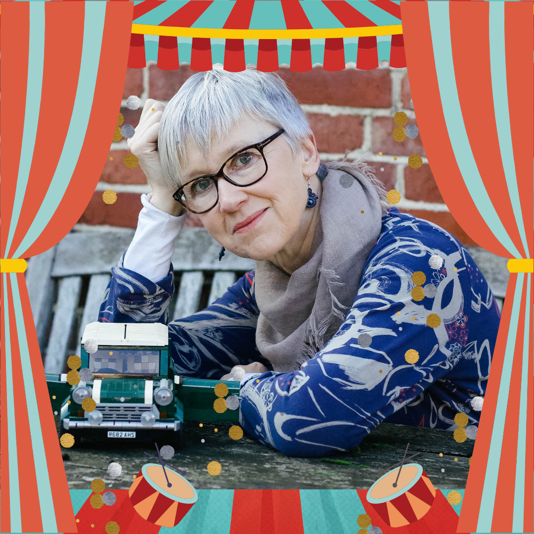 A lady with glasses looking into the camera whilst sitting leaning over a table with a lego car next to her.