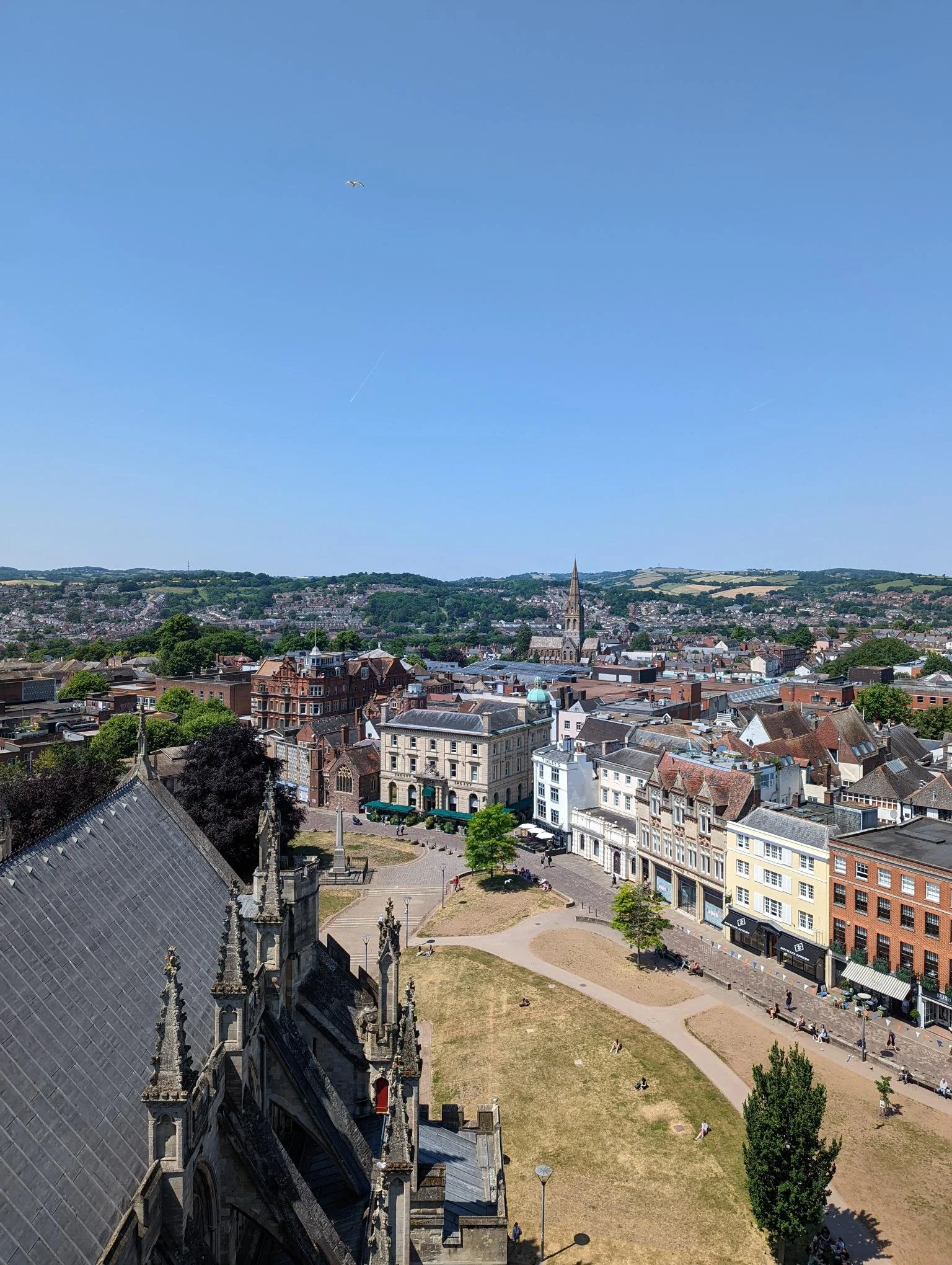 A view of a city from a cathedral roof, with cathedral turrets in the foreground, and red, white and brown city buildings in the middle ground. There are hills and fields in the background and blue skies.