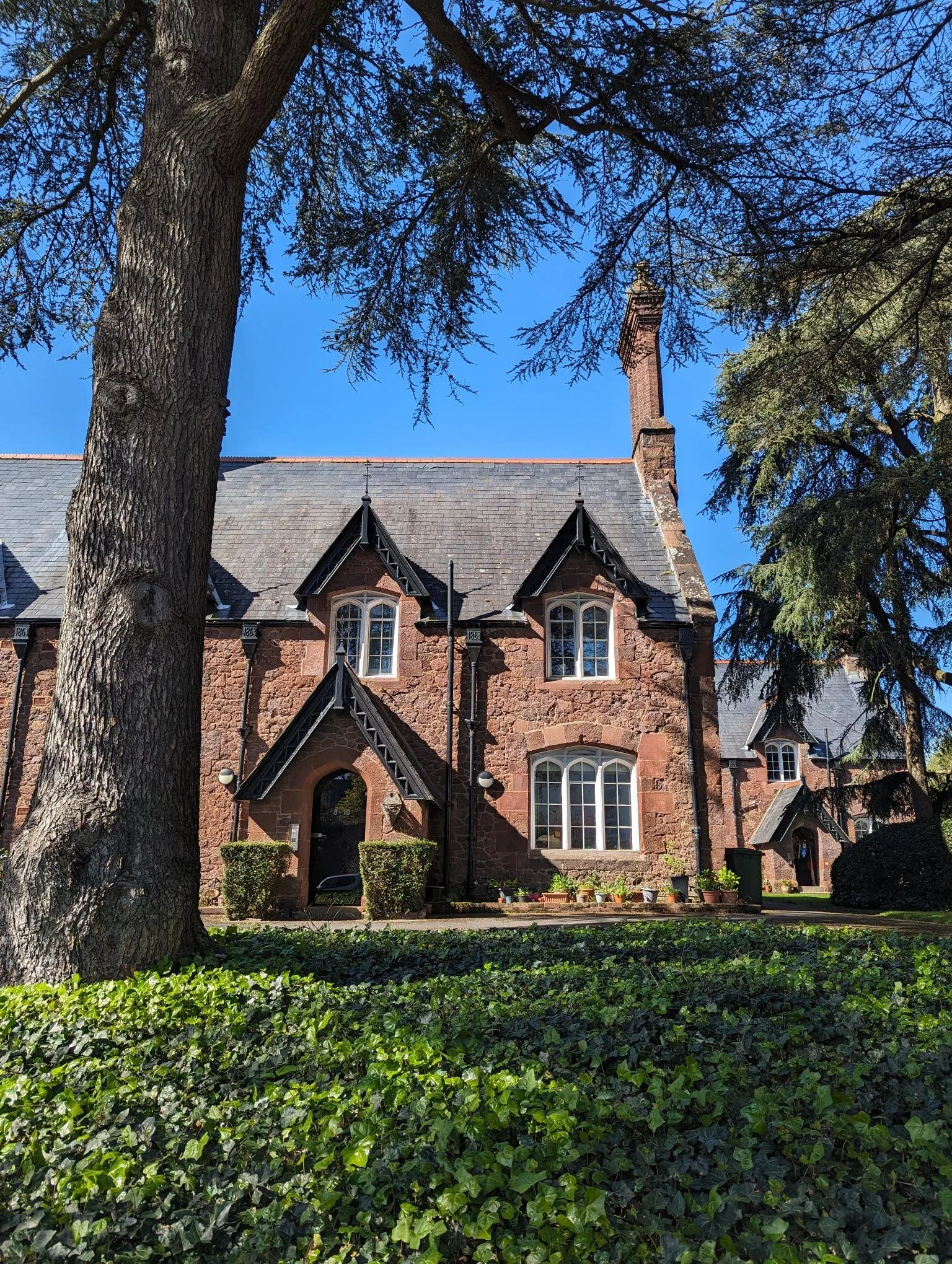 An old alms house building with red brick and pointed arches in the roof, with a tall tree and shrubbery in the foreground.