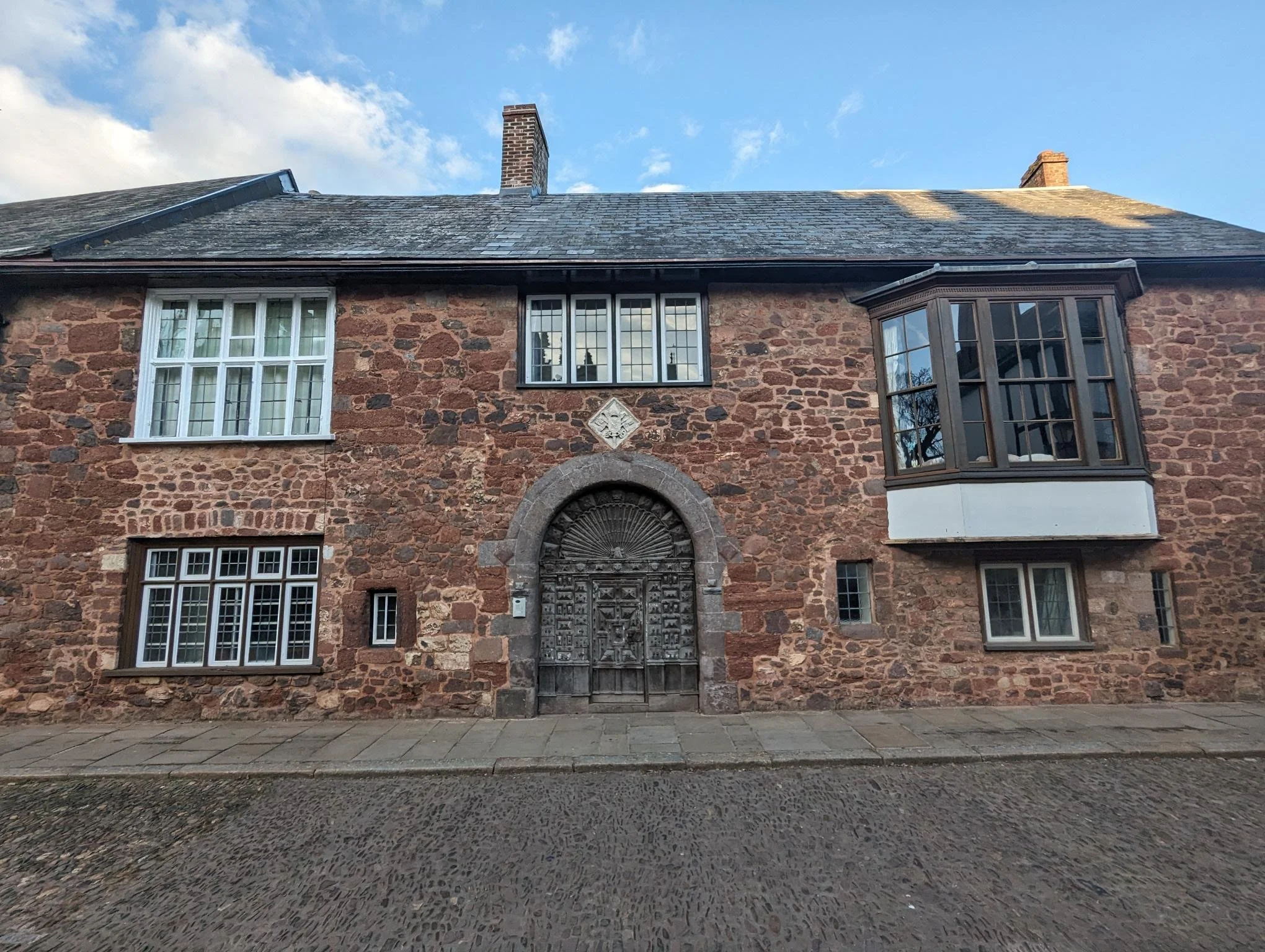 A very old red-bricked building with a studded wooden door and a crest above it, with various windows in different old styles.