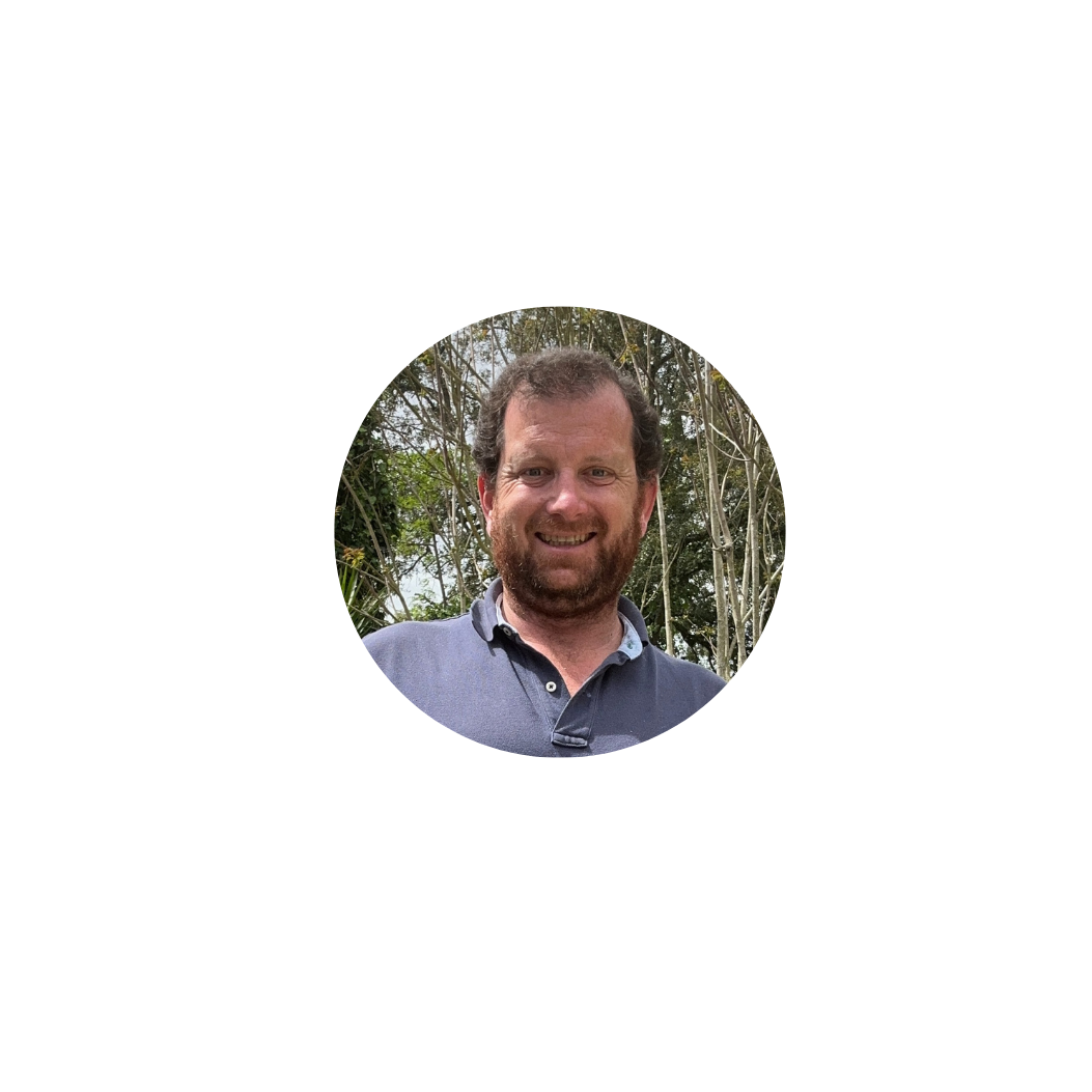 Retrato de um homem sorridente com barba e cabelo castanho, vestindo uma camisa azul, ao ar livre, com árvores ao fundo.