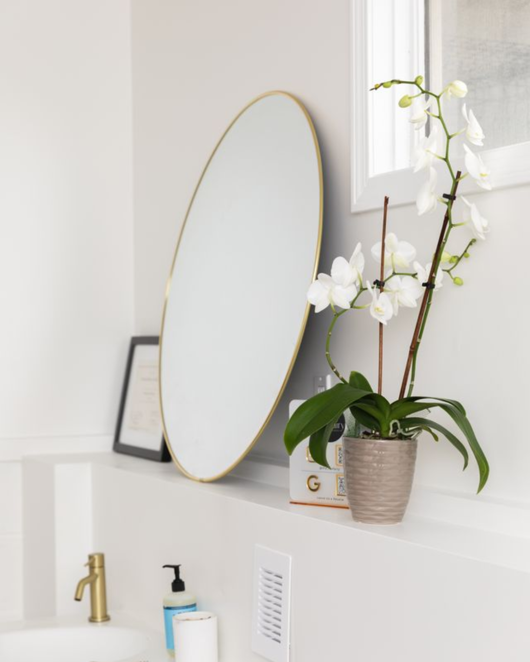 Decorative white orchid plant in a gray pot on a white ledge with a gold mirror, framed picture, soap dispenser, and a window in the background.