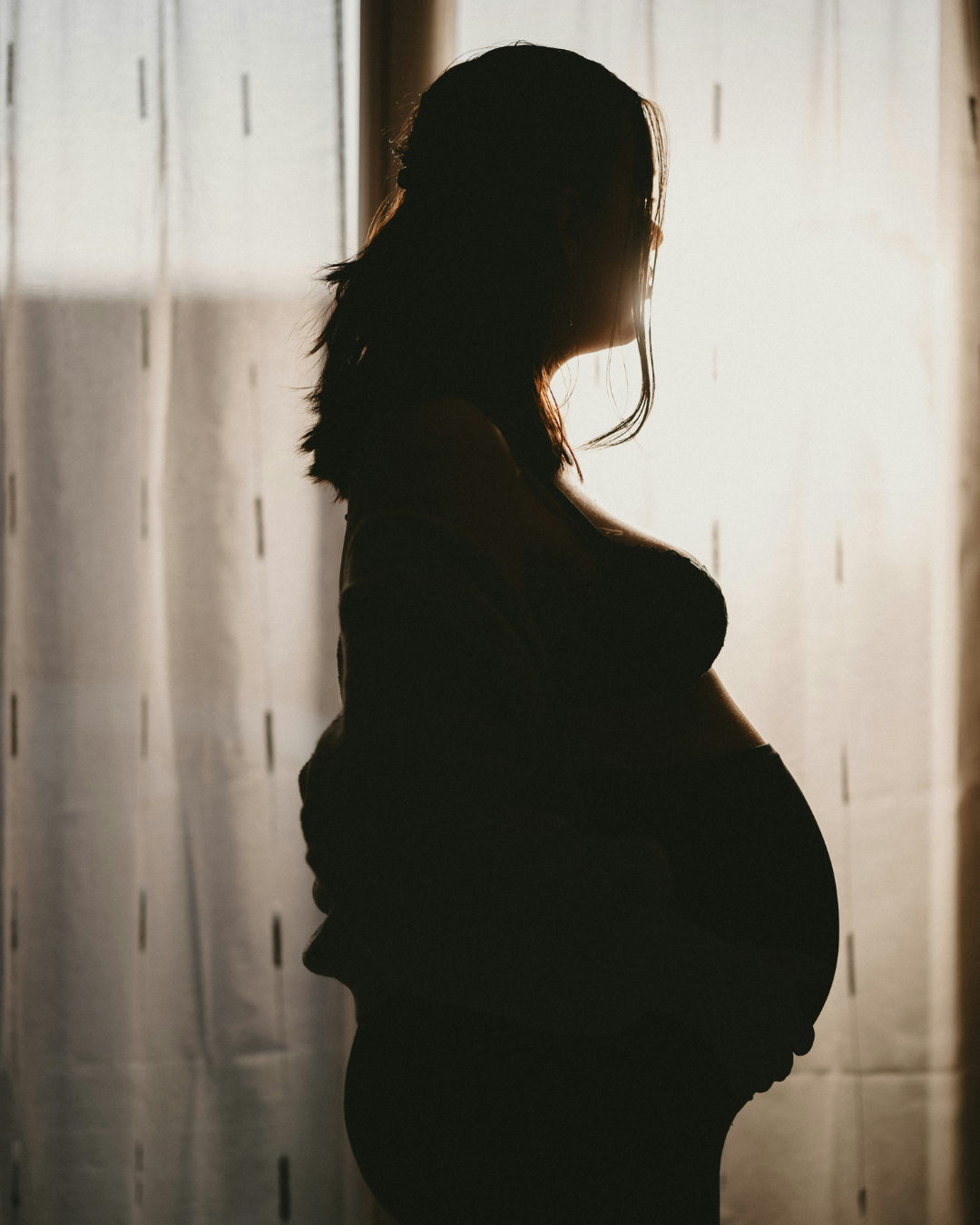 Silhouette of a pregnant woman standing near a window with backlighting.