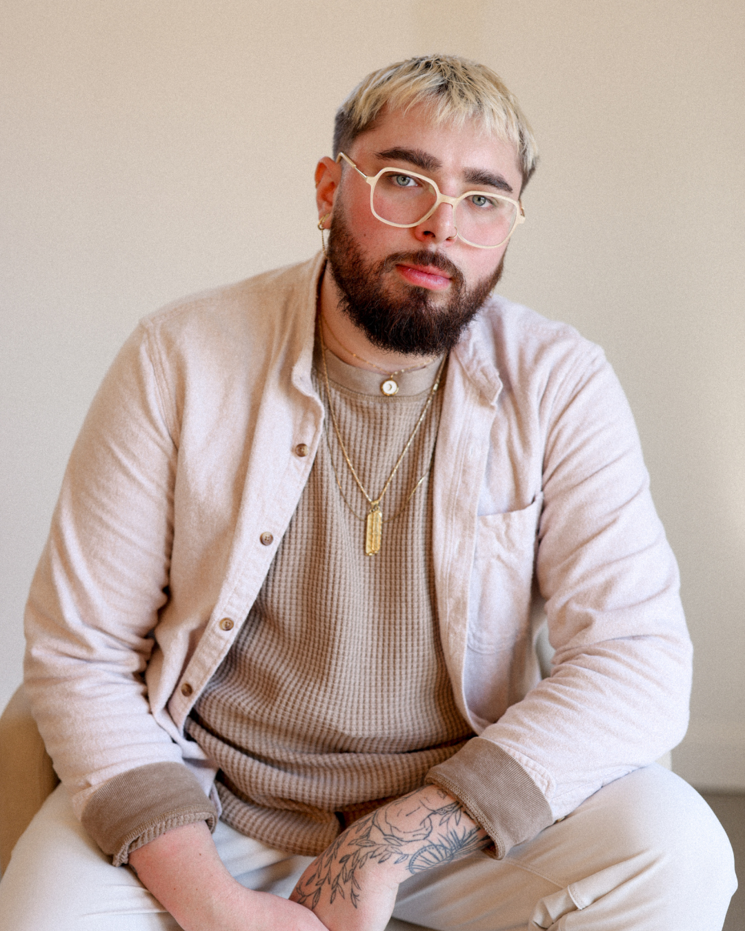 A young man with blonde and brown hair, a beard, and glasses, wearing layered necklaces, a beige button-up shirt, a beige t-shirt, and cream pants, sitting against a plain beige wall.