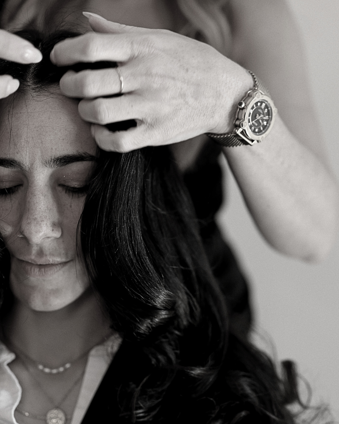 A woman with dark hair, closed eyes, and freckles, with her head bowed. An older hand with a watch is resting on her head.