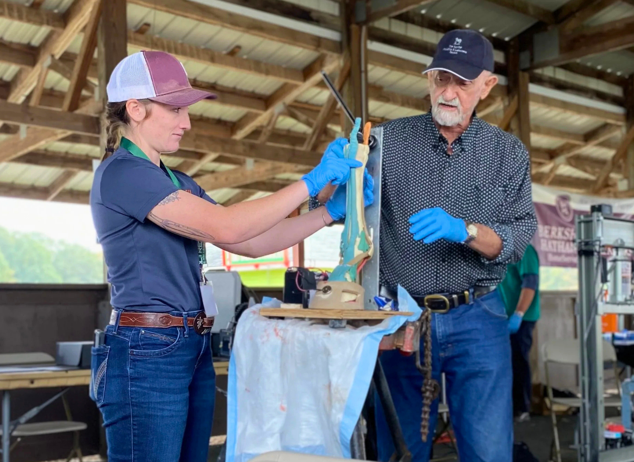 A young woman and an older man working together on a woodworking project in a barn or workshop. The woman, wearing a baseball cap, t-shirt, and gloves, is holding a saw or similar tool, while the older man, wearing a cap and patterned shirt, is assisting. Snowy or cloudy outdoor scenery is visible through the open sides of the barn.