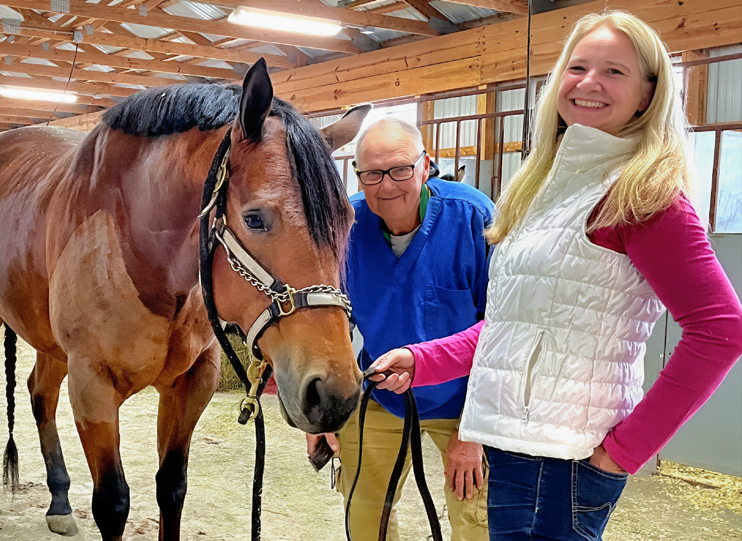 A smiling young woman with long blonde hair standing next to a horse in a barn, with an older man wearing glasses and a blue jacket behind them.