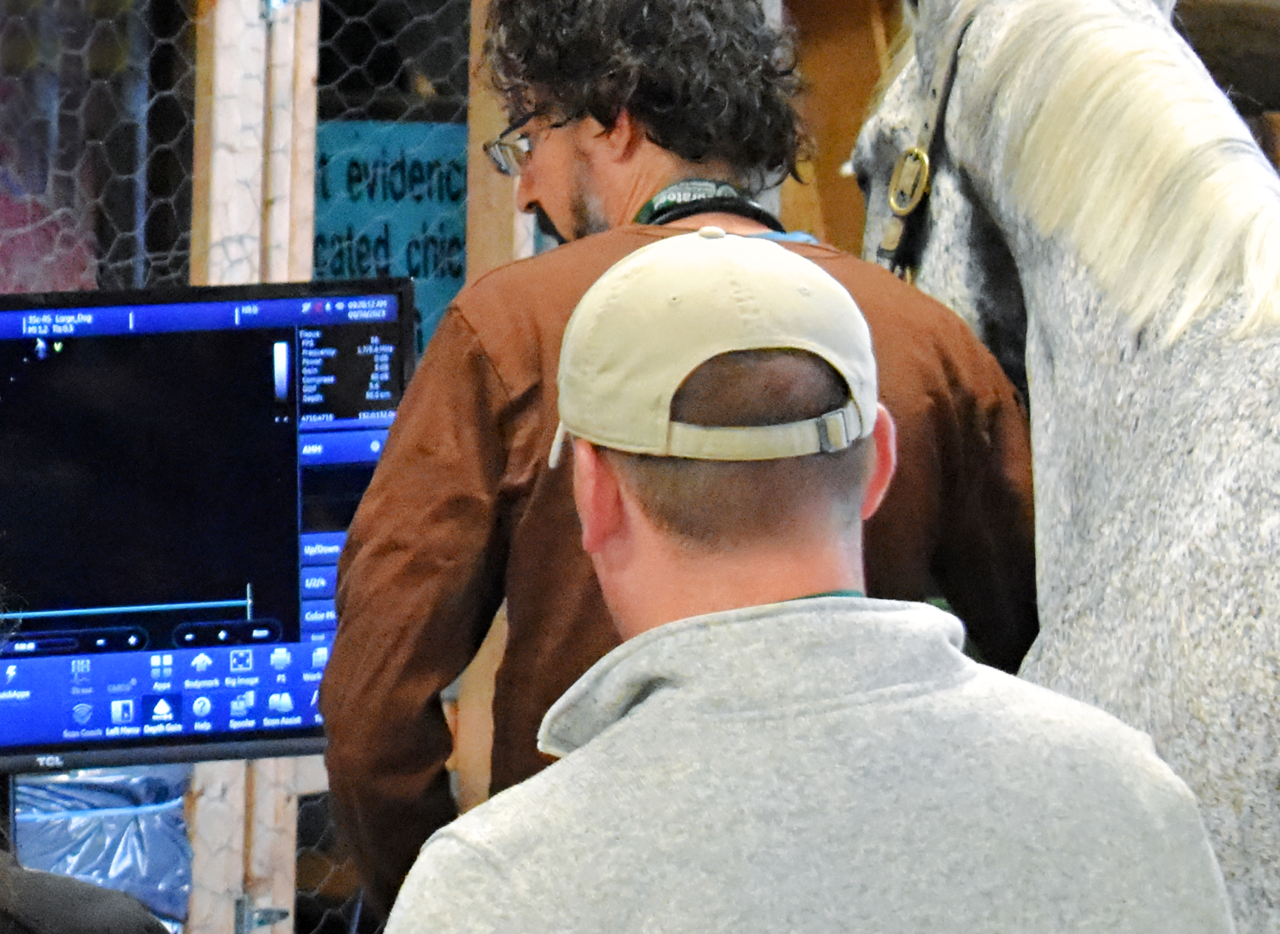 Two men, one with curly dark hair and glasses, and the other with a backwards baseball cap, are standing in front of a computer monitor. The background includes a wooden frame and a chain-link fence, suggesting they might be in a workshop or construction site. The man with glasses is wearing a necklace and a brown jacket, while the other is wearing a grey hoodie.