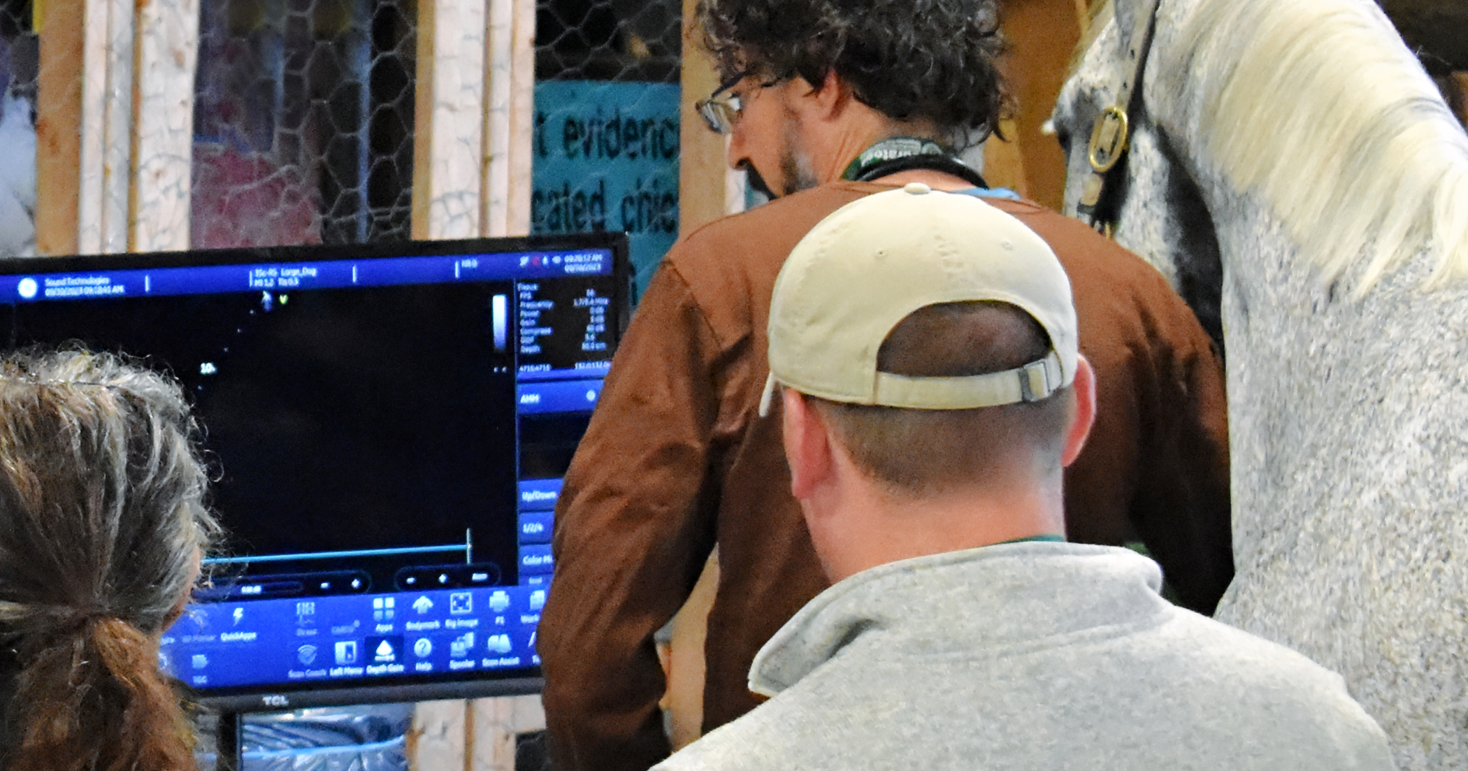 Three people gathered around a computer monitor showing a software interface, with a piece of wood and wooden framing in the background.