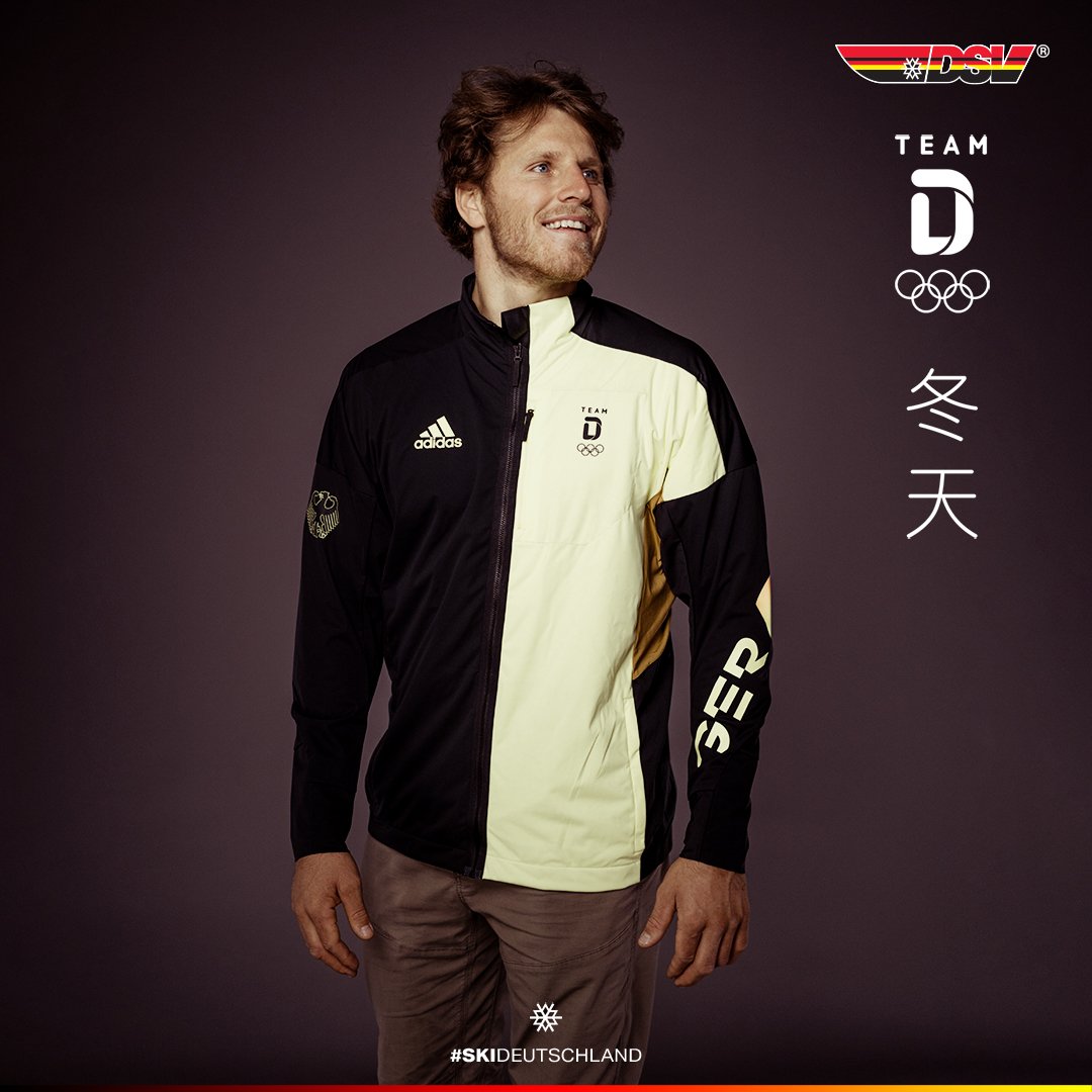 A person wearing a black and yellow Team Germany ski jacket with Adidas branding and Olympic rings, standing against a dark background. The jacket has 'GER' text on the sleeve. The image has logos, Chinese characters, and the hashtag #SKIDEUTSCHLAND