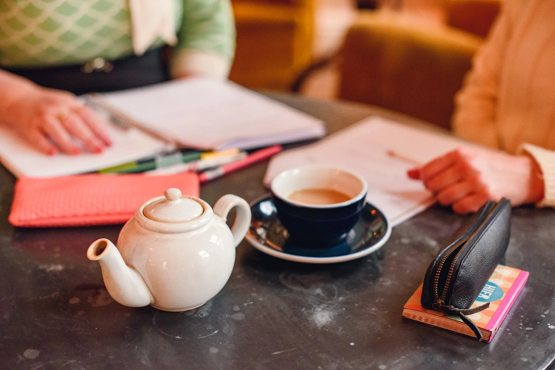 A tea setting on a black table with a white teapot and a cup of tea, surrounded by notebooks, pens, and a black pencil case, with hands gesturing and a person sitting nearby.
