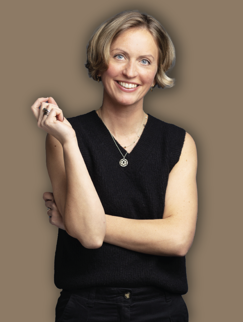 A woman with short blonde hair and blue eyes smiling, wearing a black sleeveless top, jewelry, and dark pants, standing against a brown background.