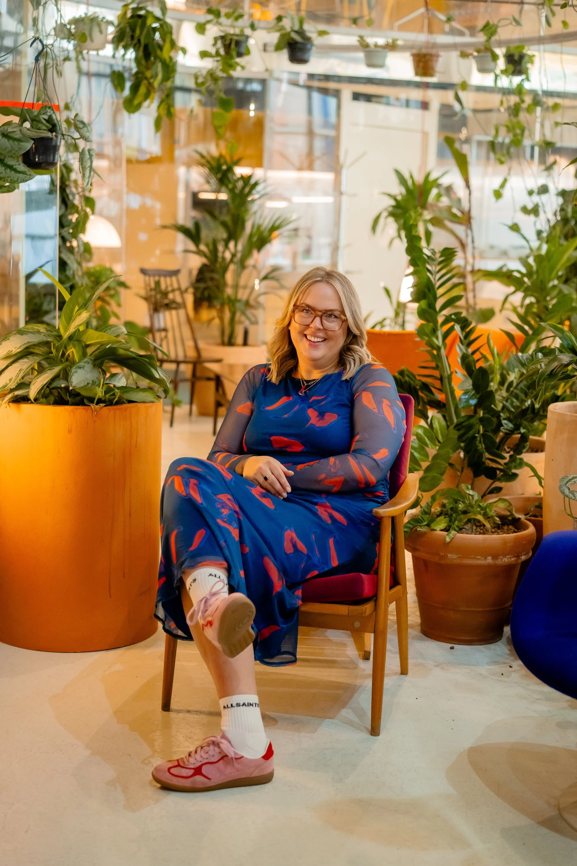 A woman with blonde hair, glasses, and a blue dress with red patterns is sitting on a wooden chair surrounded by various potted plants in a bright, modern indoor space. She is smiling and appears relaxed.