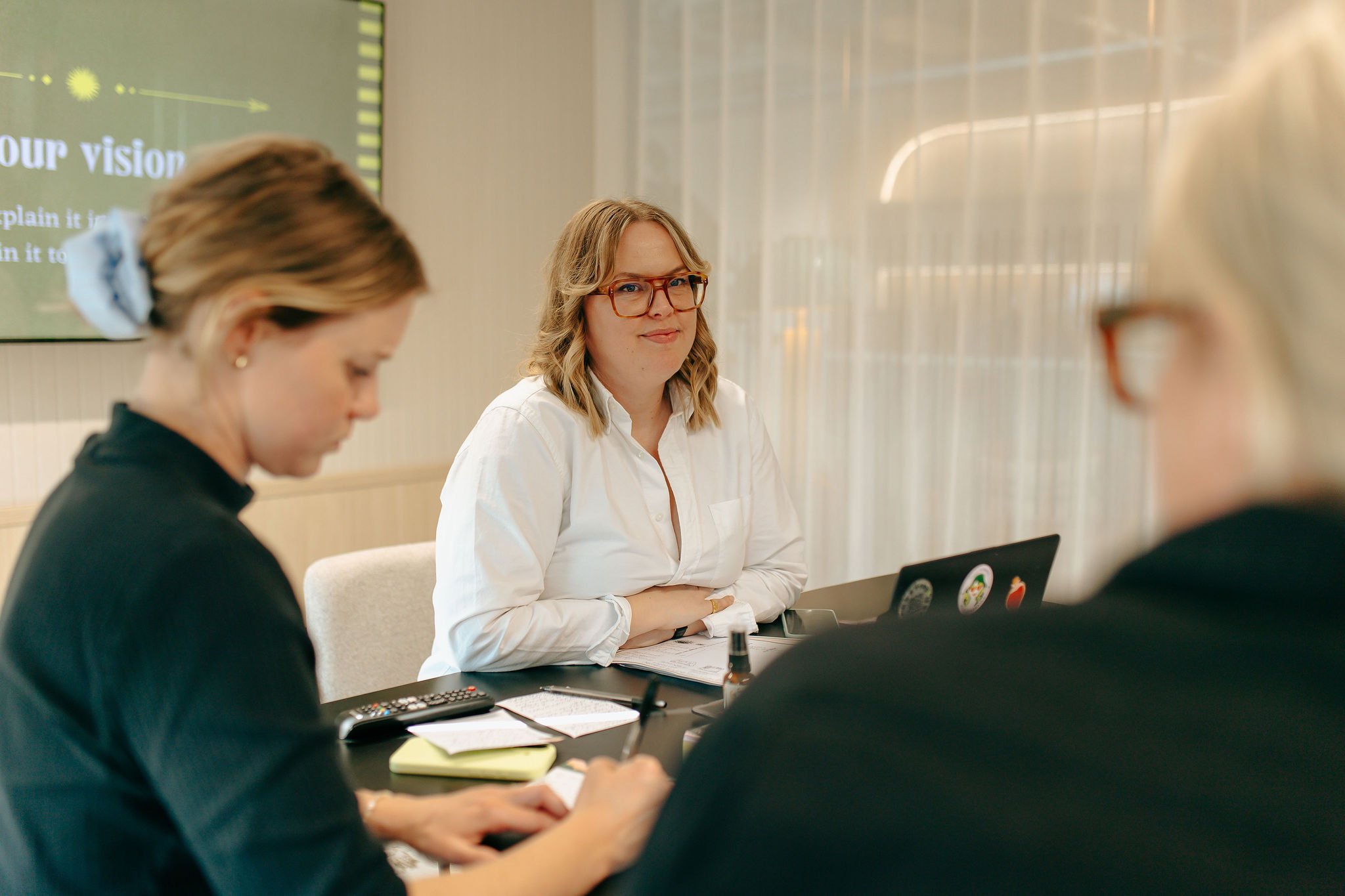A woman with blonde hair and glasses sits at a table during a meeting, with other people partially visible in the foreground. Behind her is a screen displaying a presentation.
