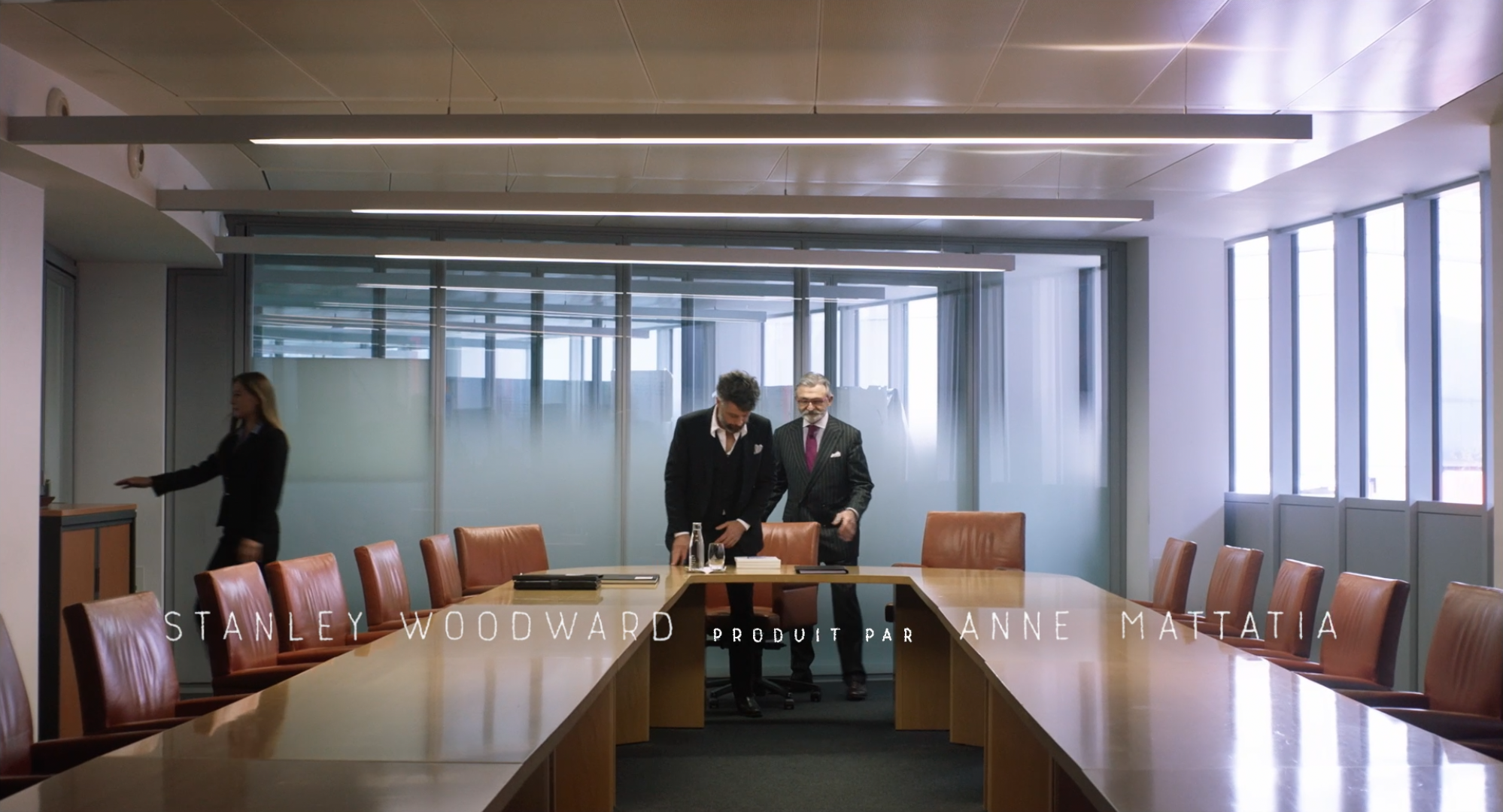 Salle de réunion avec quatre personnes, deux hommes en costume au centre, une femme à gauche, grand tableau de verre à l'arrière, tables en U avec chaises en cuir marron.