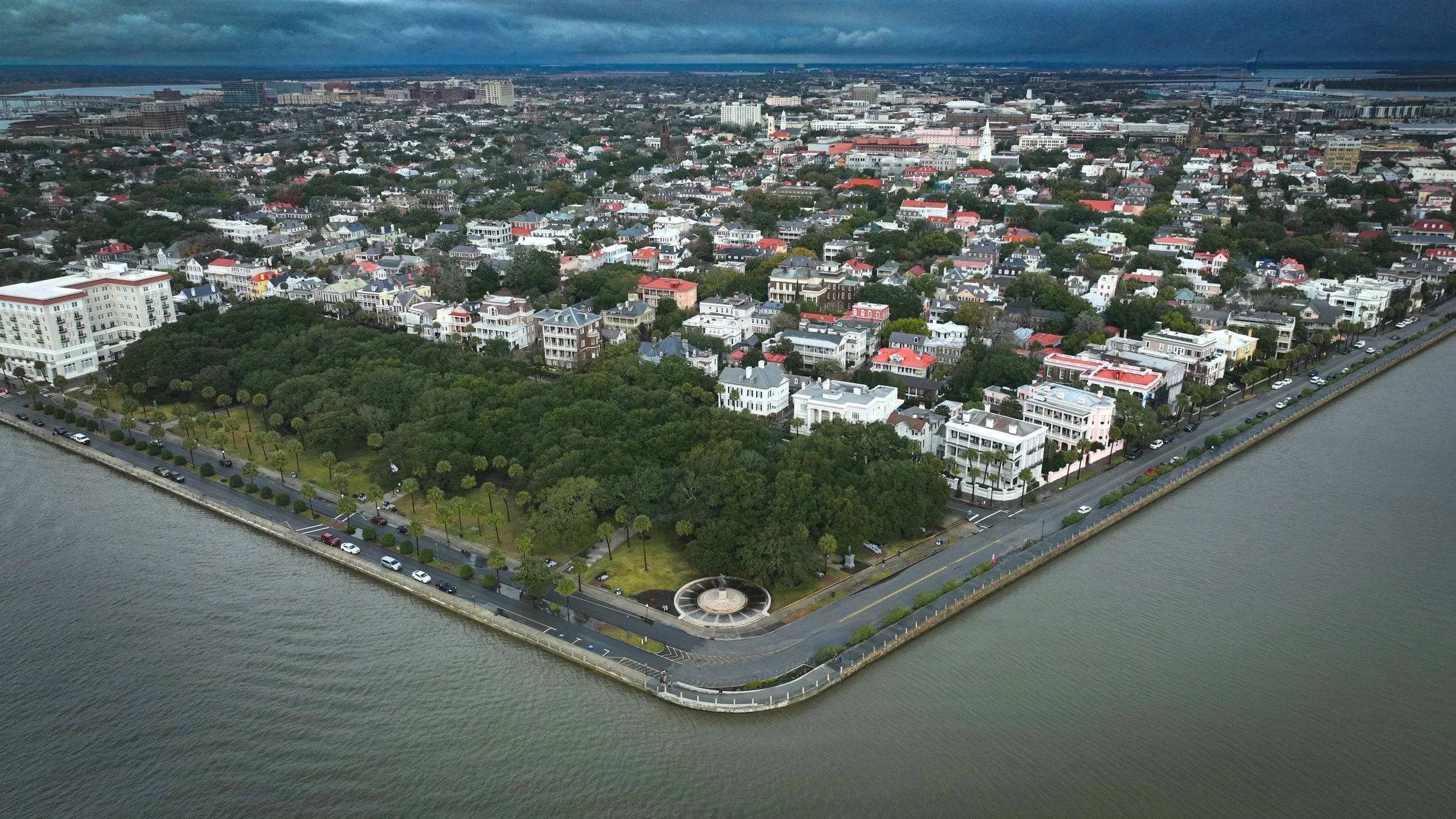 Aerial view of a city with a park and waterway, featuring numerous colorful houses and buildings, and cloudy skies overhead.