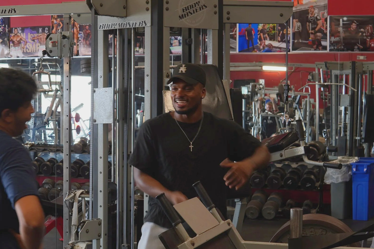 A muscular man in a gray tank top taking a selfie in a gym with various fitness equipment in the background.