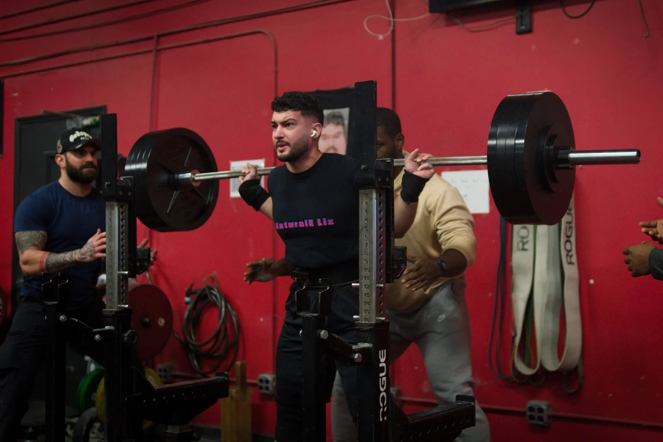 A man with curly hair and a beard performing a squat lift with a loaded barbell at a gym, surrounded by other people.
