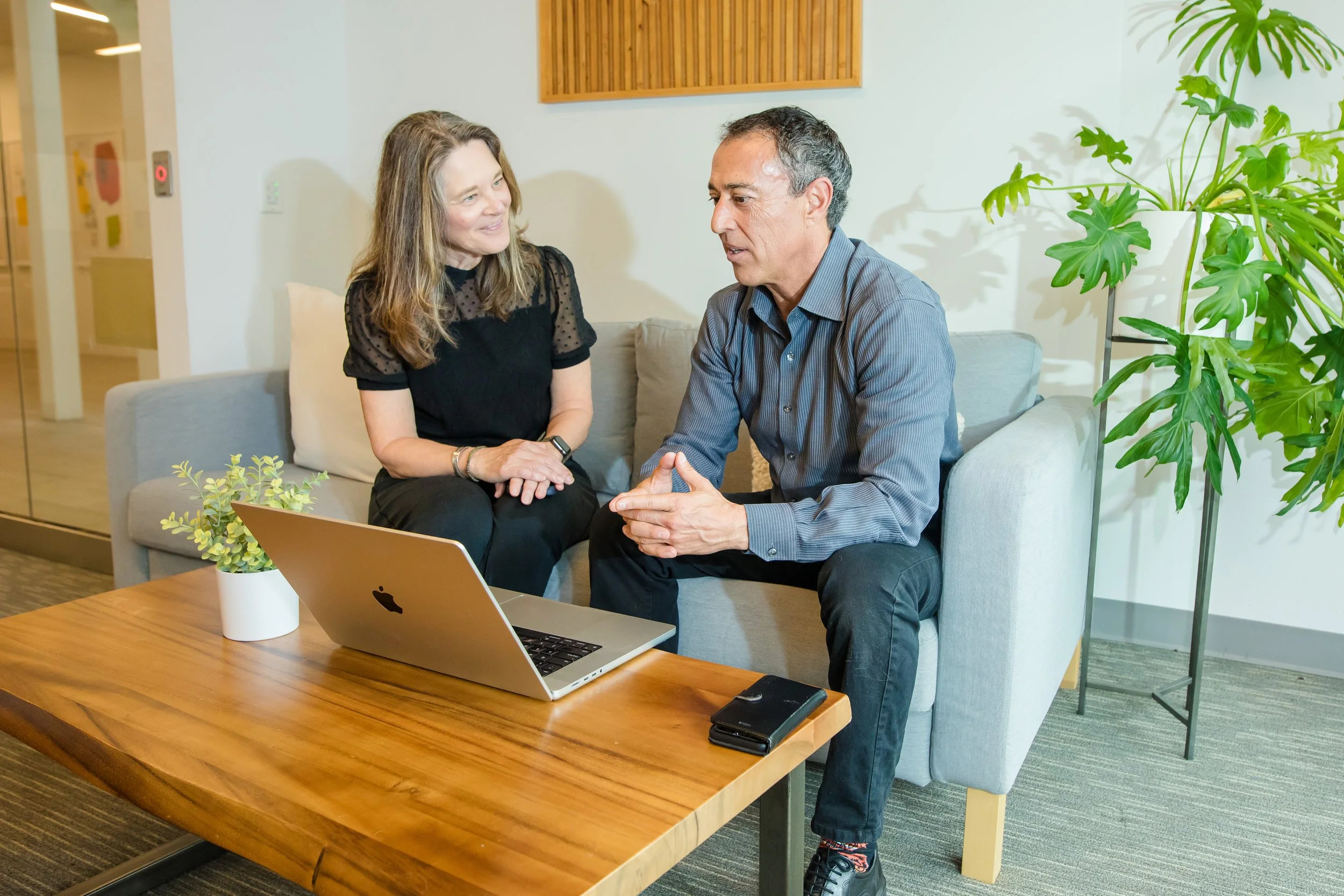 A man and woman sitting on a light gray couch in a modern office, engaging in a conversation with a MacBook open on a wooden coffee table in front of them.