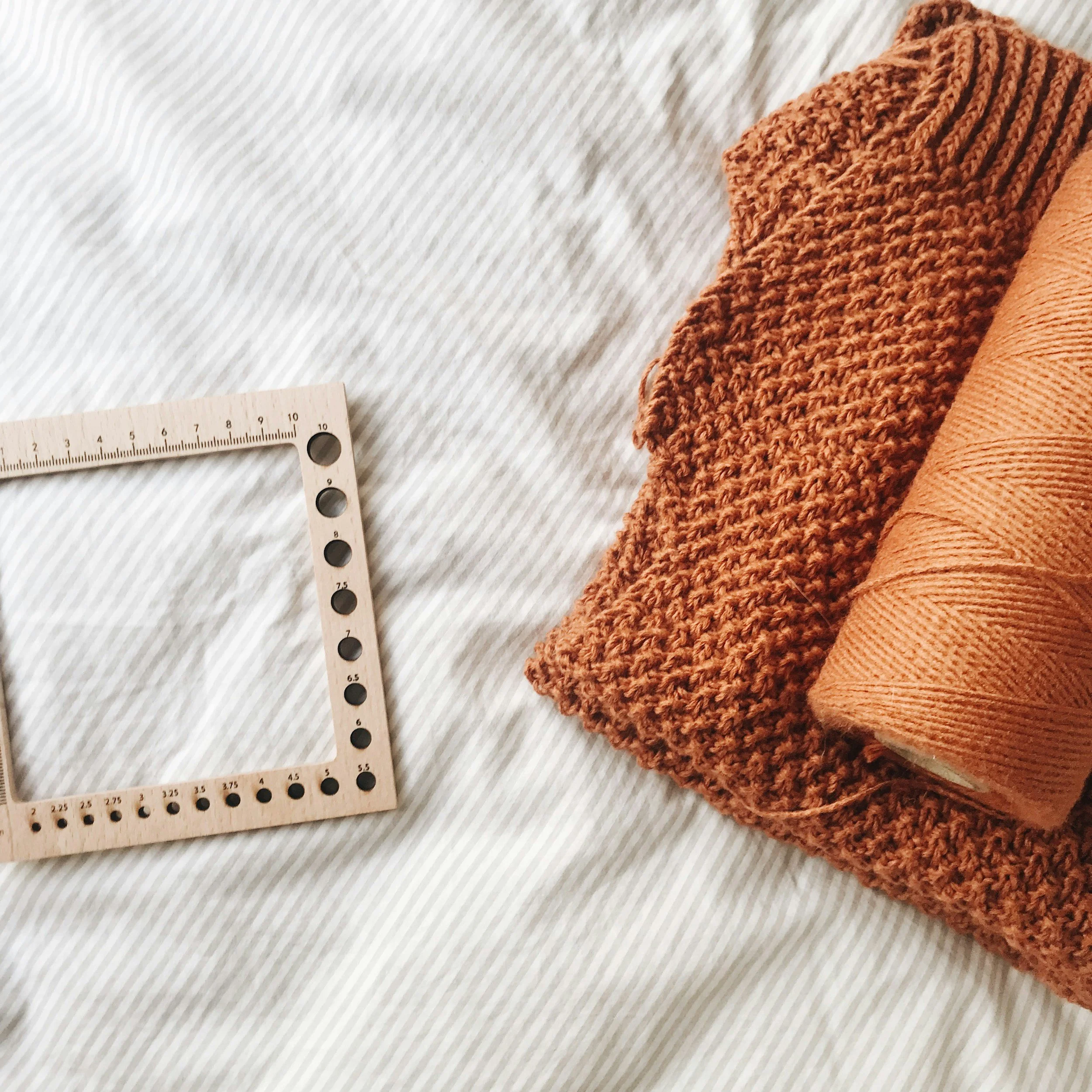 Rust-orange seed stitch knit swatch with a wooden gauge ruler and matching yarn spool on striped fabric