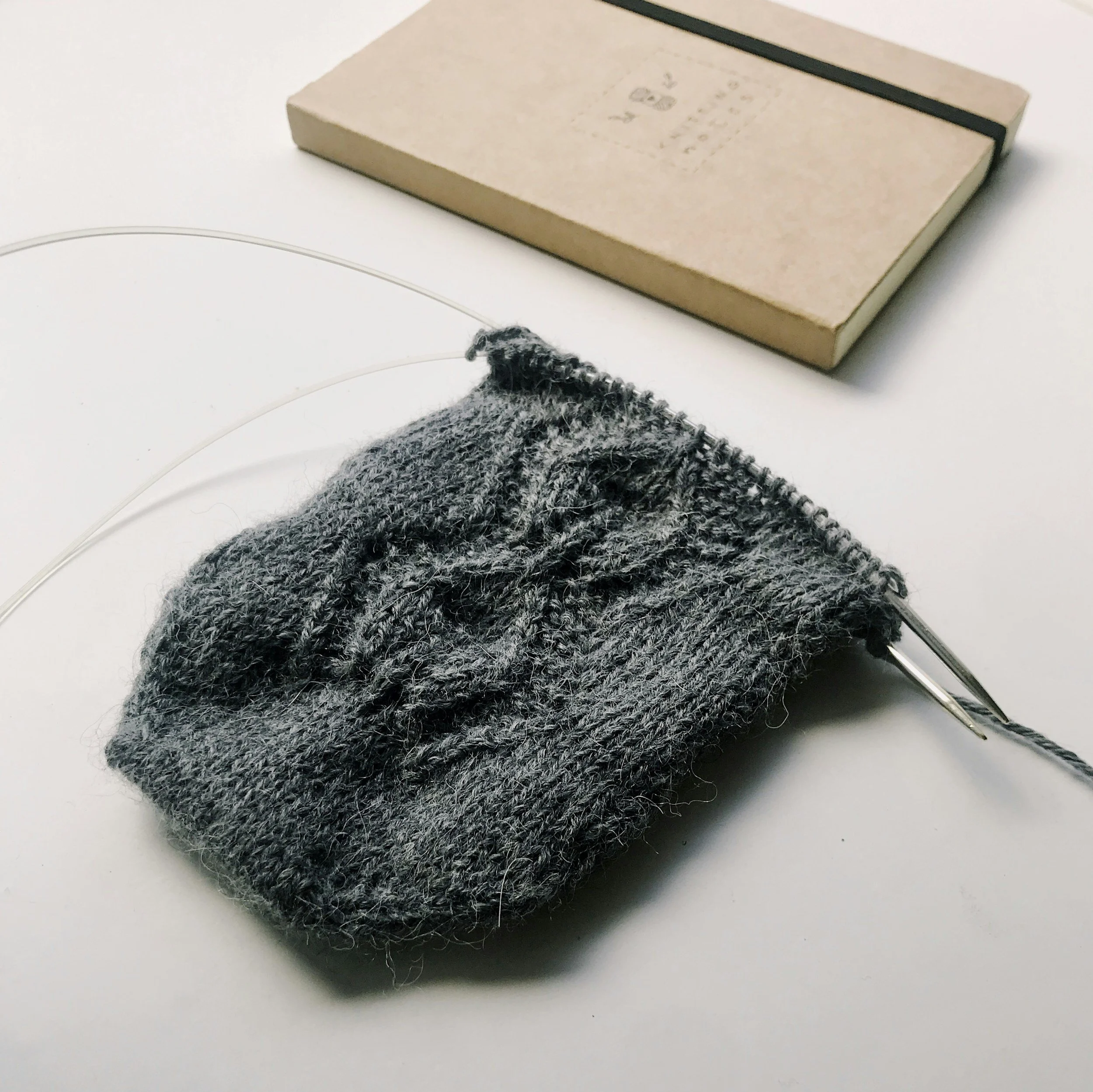 Gray cable knit swatch in progress on circular needles, resting on a tan notebook