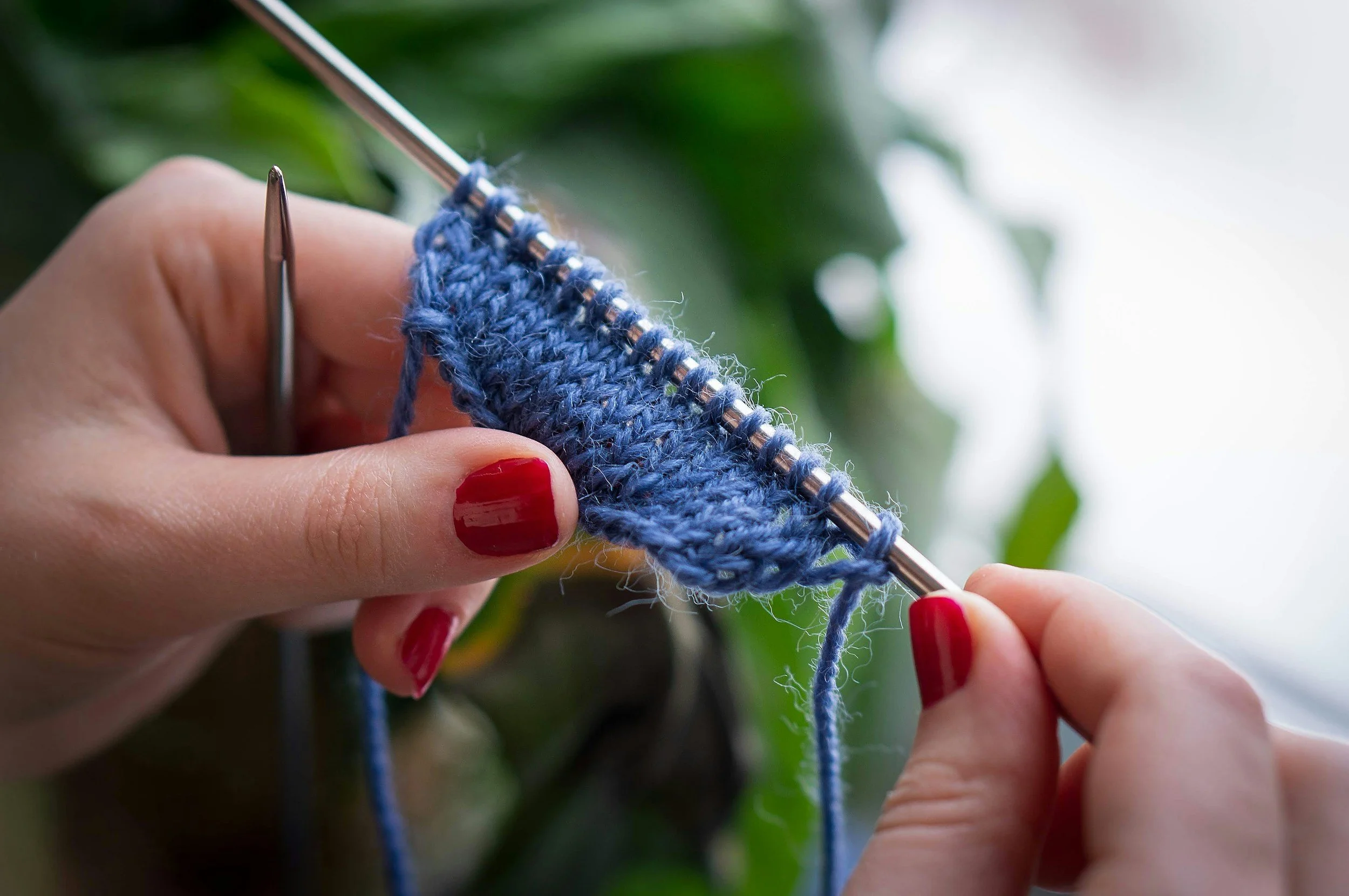 Close-up of hands with red nail polish knitting blue yarn on metal needles, greenery in the background