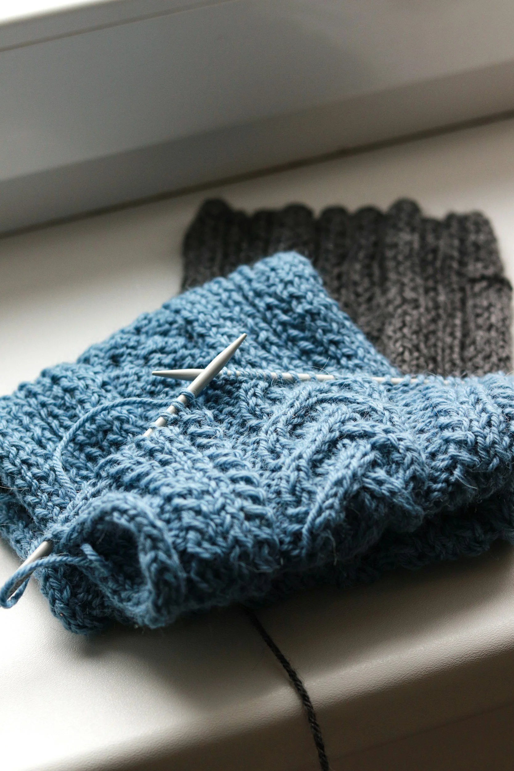 Blue ribbed knit fabric on circular needles resting on a windowsill, with a dark gray knit piece folded behind it