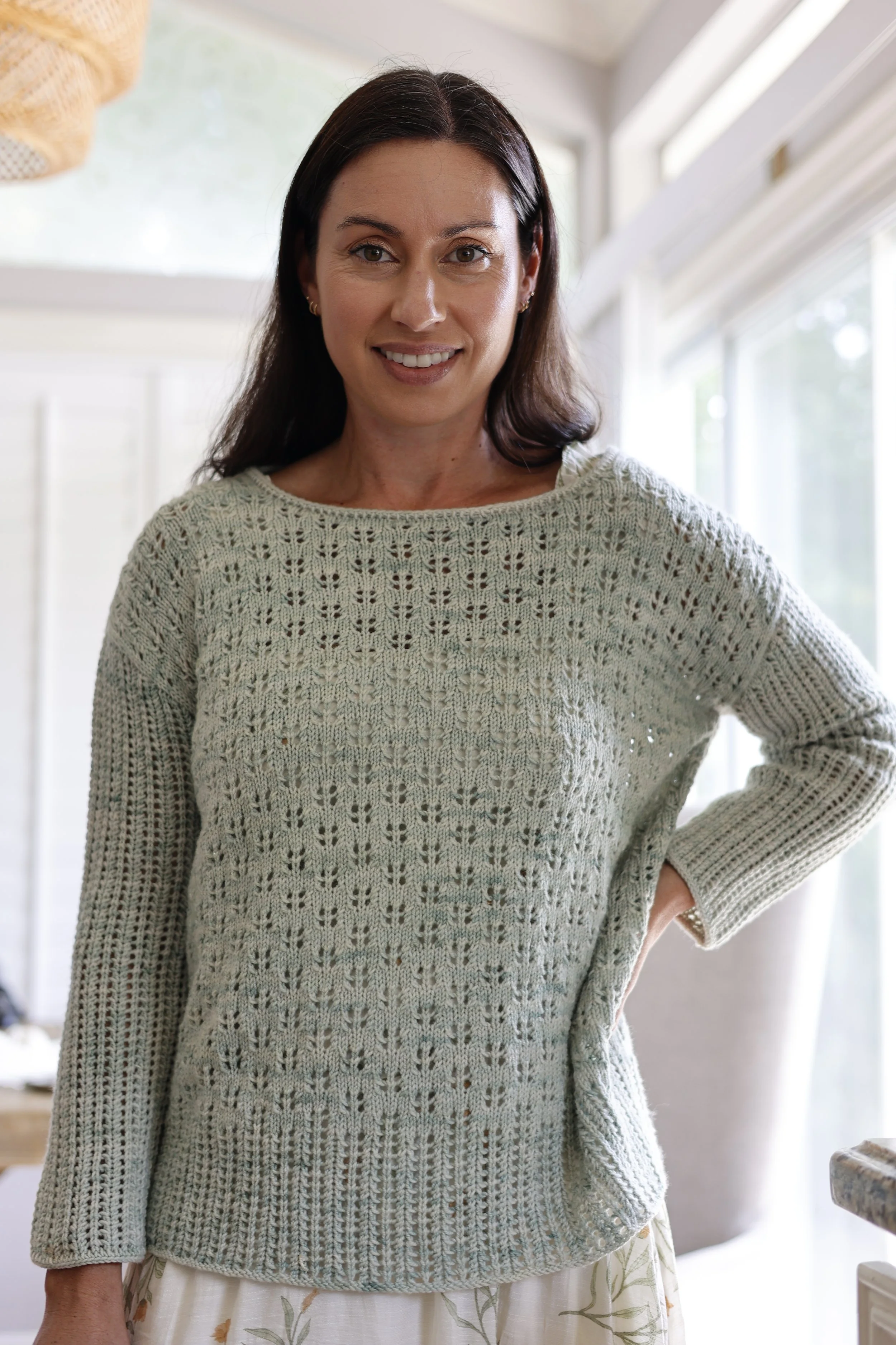 Woman with dark hair wearing a sage green eyelet lace knit pullover with a boatneck neckline and long sleeves, standing indoors near a window