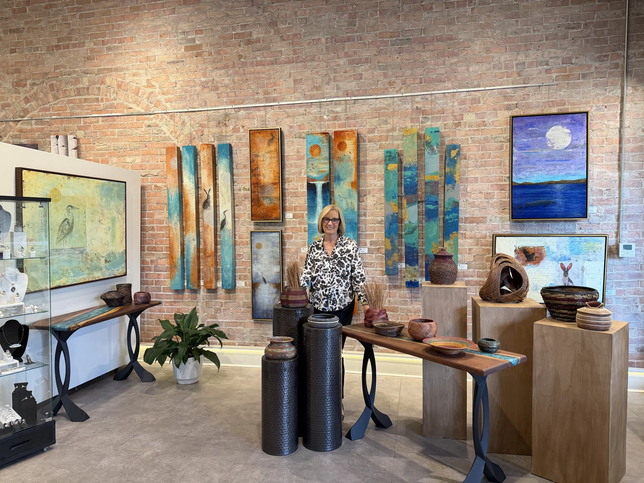Interior of an art gallery with paintings on exposed brick wall, a woman in a black and white patterned shirt standing in front, and various art pieces and displaying furniture around.