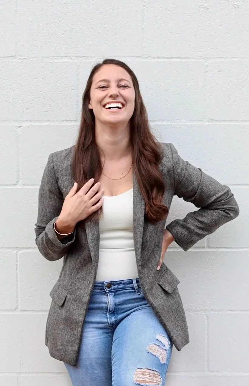 Woman with long brown hair is smiling and laughing, wearing a gray blazer, white top, and ripped jeans, standing against a white brick wall.