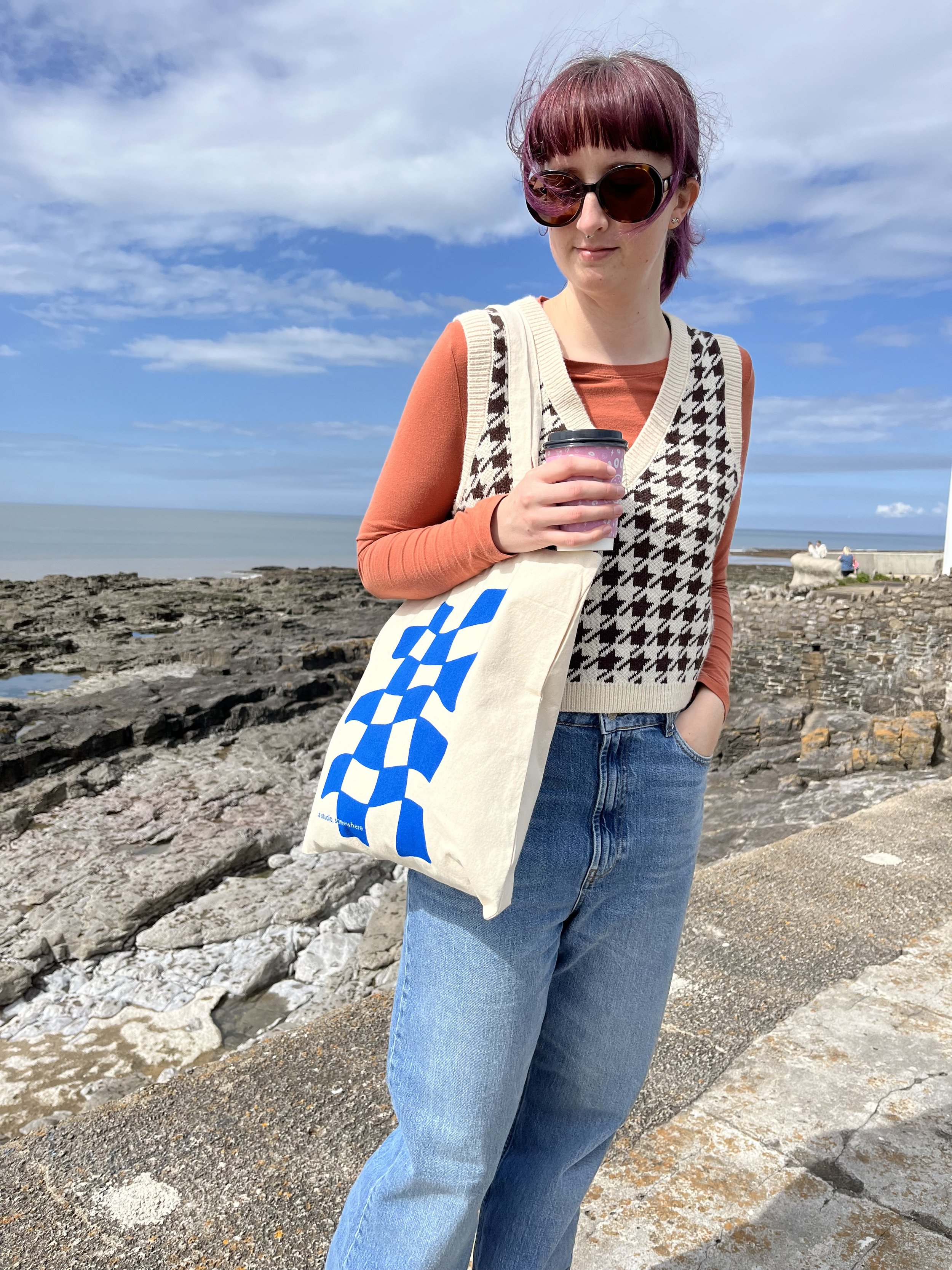 Woman holding tote bag and a coffee cup, next to the sea
