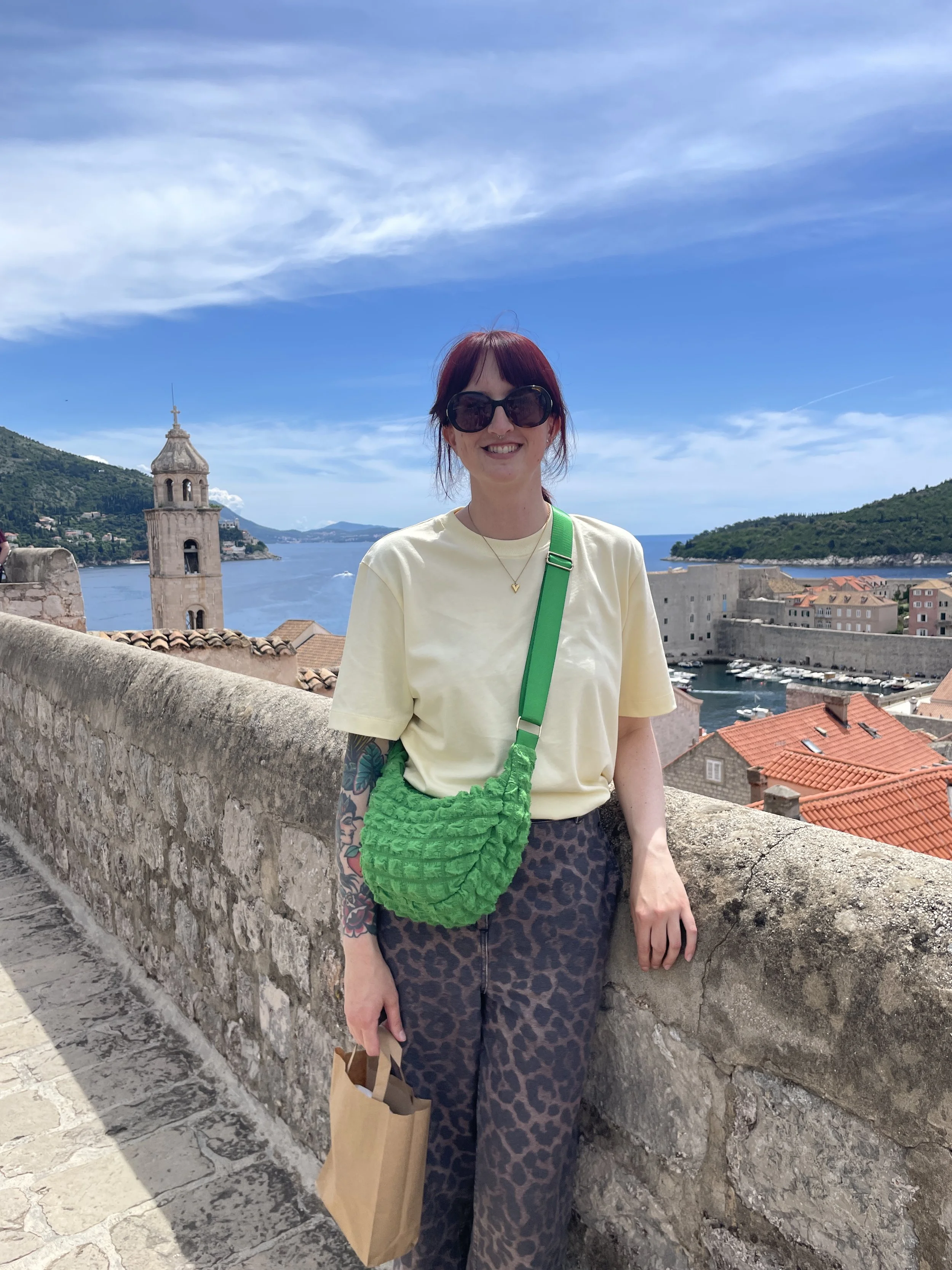 A woman with red hair and dark sunglasses smiling on a historic stone bridge, with a scenic coastal town and blue water in the background.