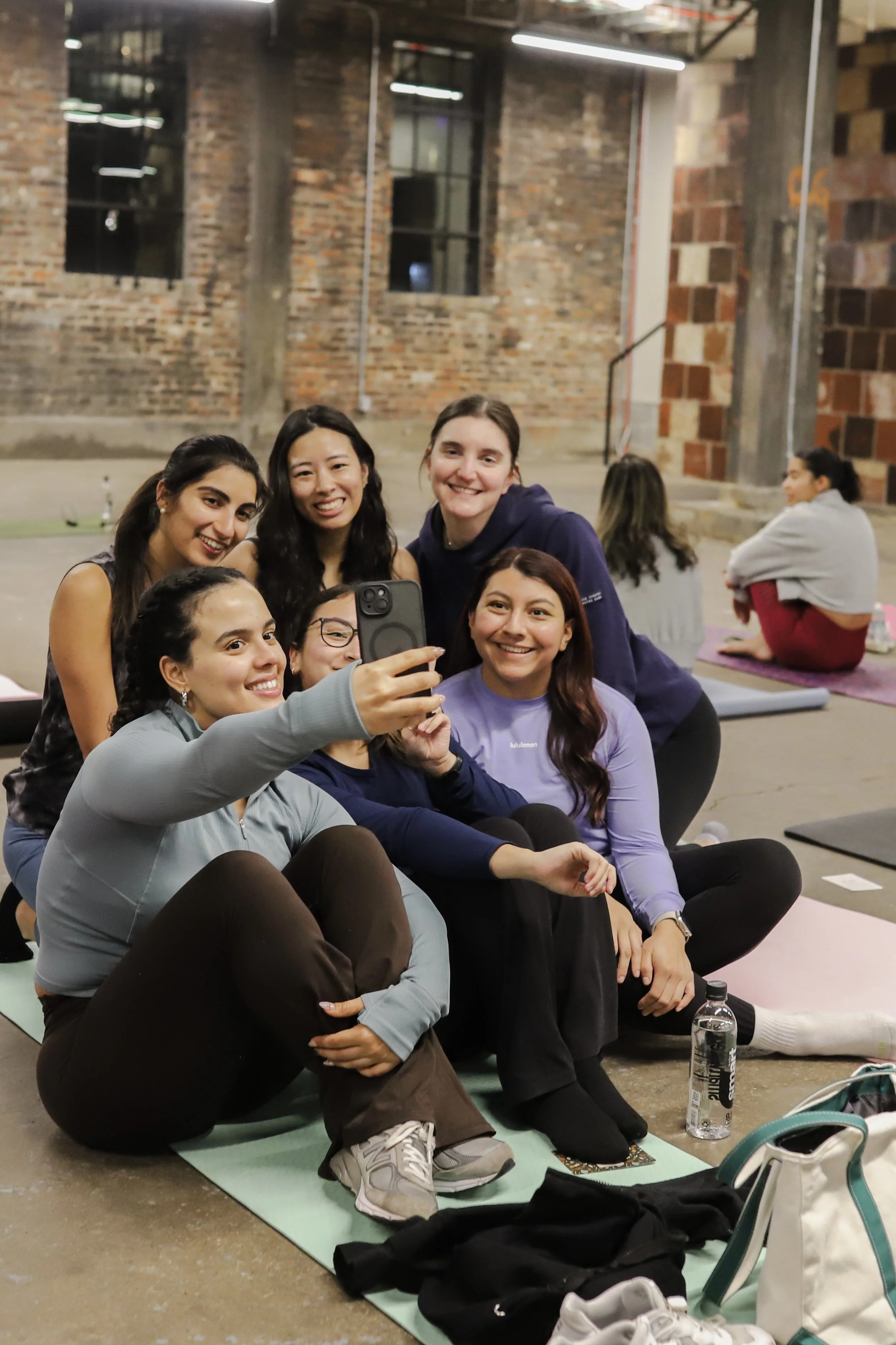 Group of six women taking a selfie together in a gym or yoga studio with exposed brick walls, other women practicing yoga in the background, and yoga mats on the floor.