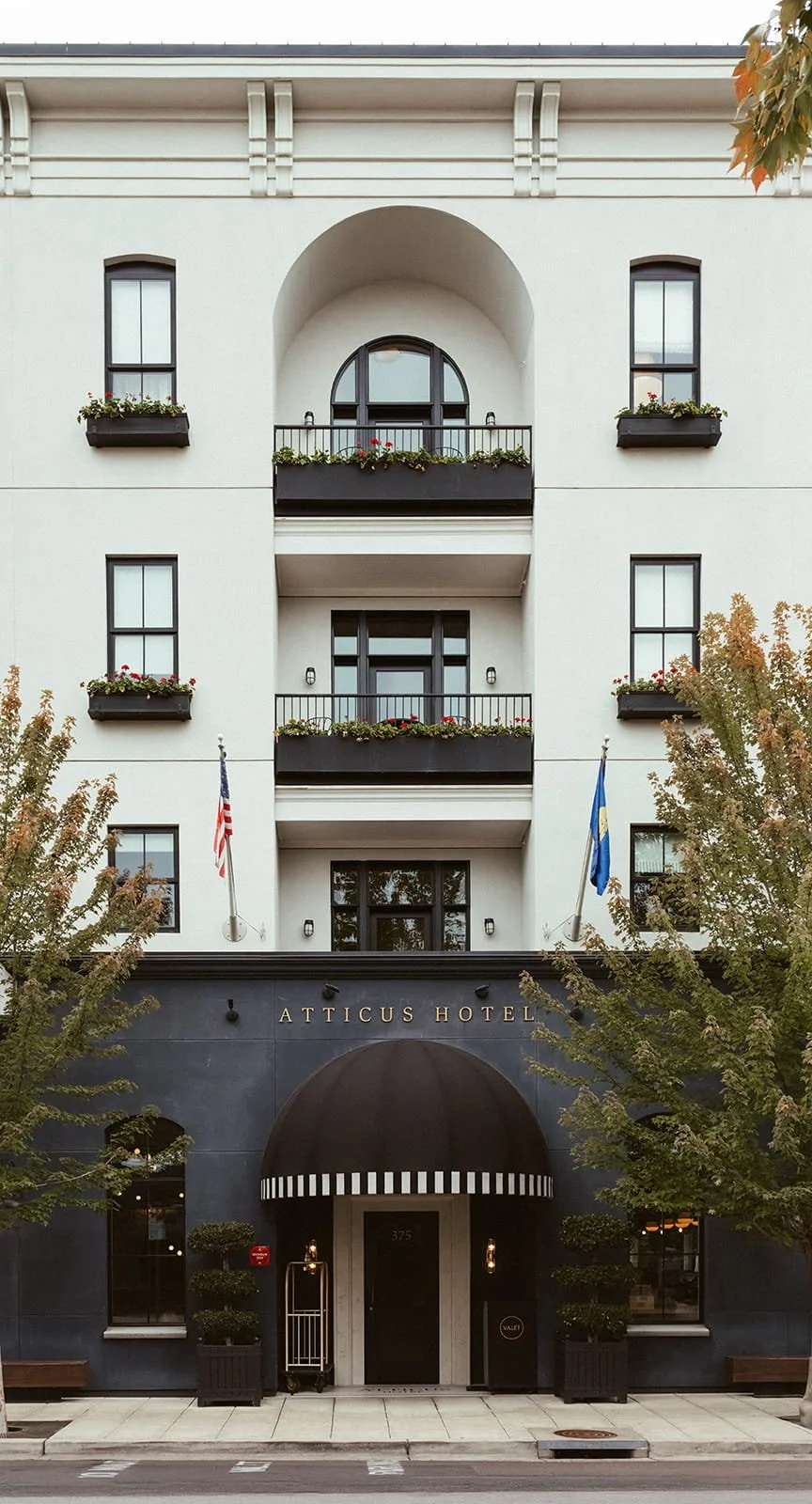 Front view of the Atticus Hotel, a multi-story building with white walls, black balconies with flower boxes, and a dark awning over the entrance. Flags are displayed on either side of the entrance, and trees frame the scene.