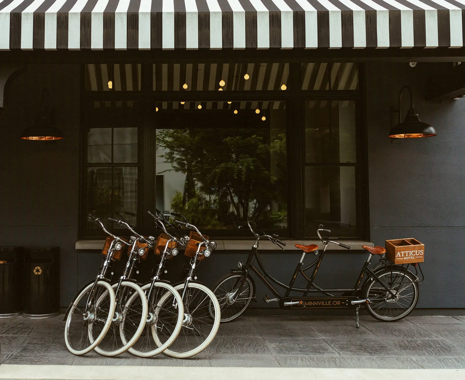 A row of five white bicycles lined up in front of a dark building with a window and black walls. There is a black bicycle with a wooden crate on the back, labeled "ATTICUS HOTEL."