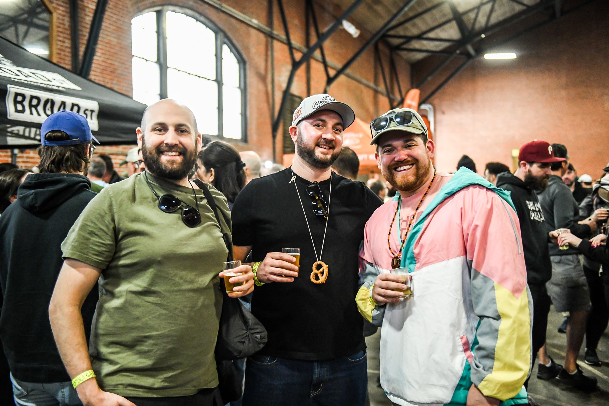 Three men smile at the camera at an indoor tasting event, each holding a small drink, with a lively crowd and vendor booths behind them.