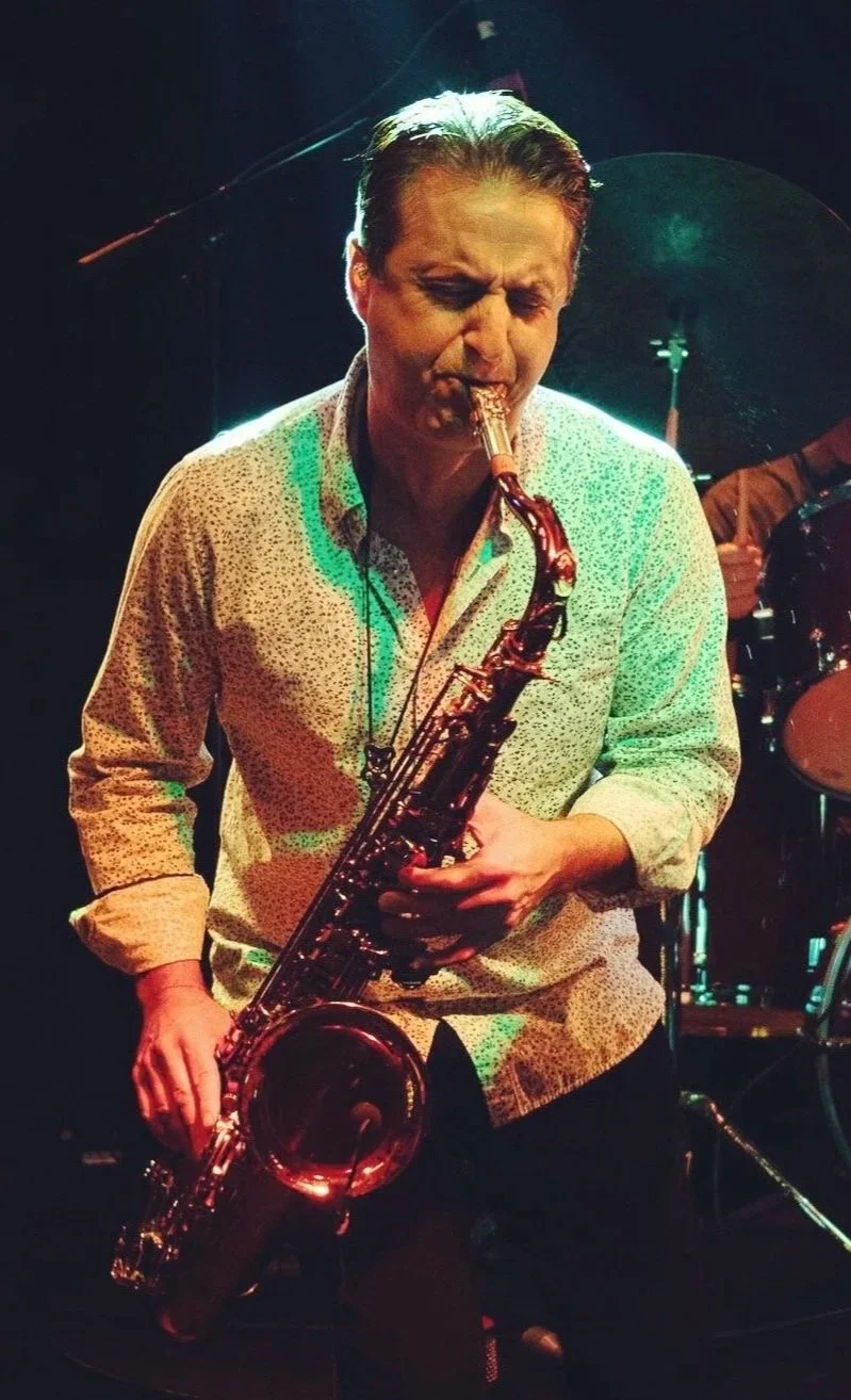 A man playing the saxophone on stage with a drum set in the background.