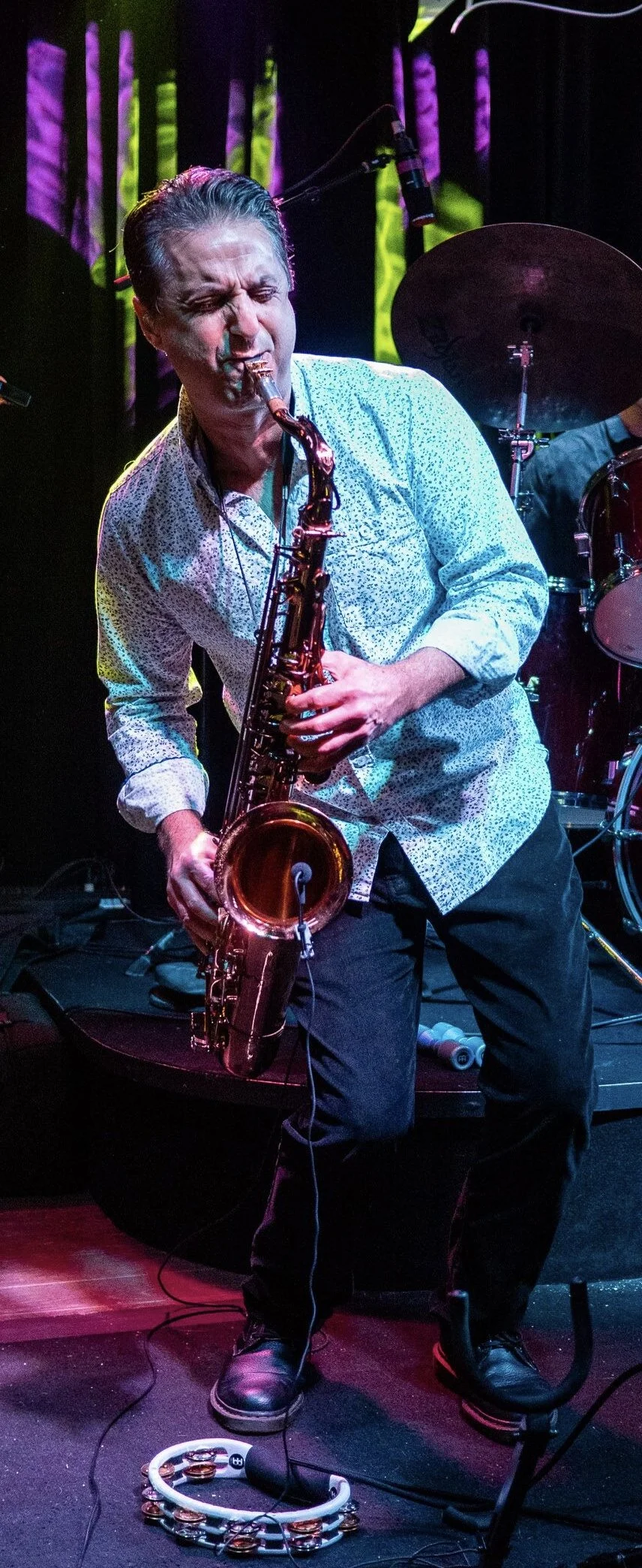 Jazz musician playing the saxophone on stage with drums and colorful lighting in background.
