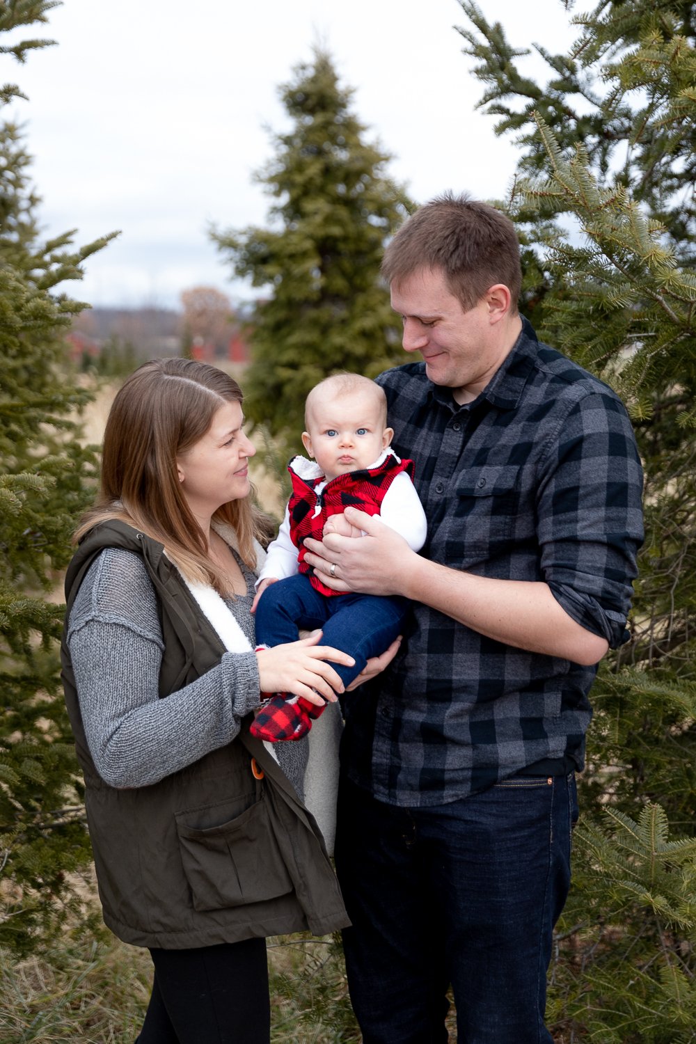 mom and dad holding their daughter in front of an evergreen tree at Poplar creek tree farm
