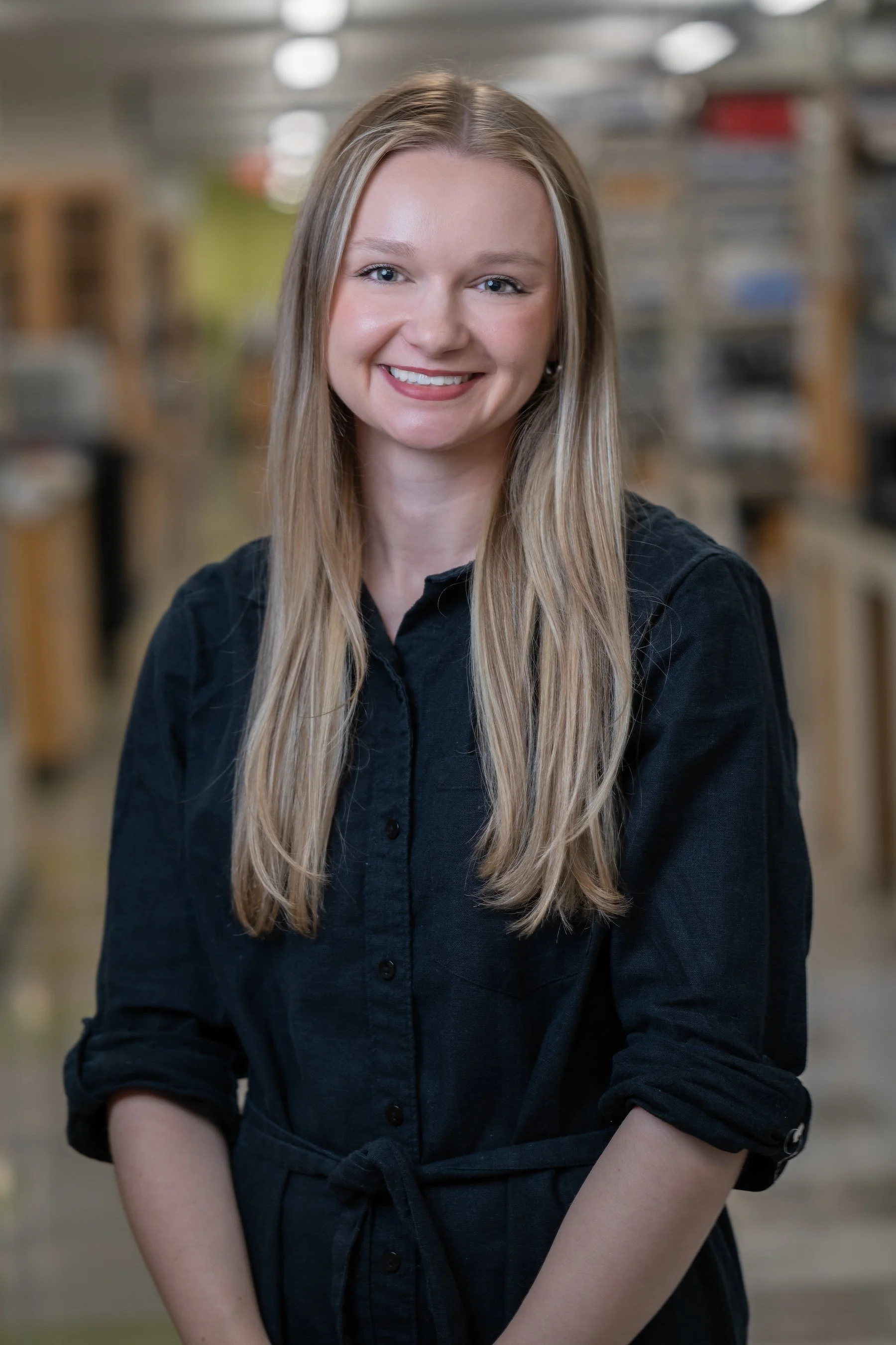 A young woman with long blonde hair smiling, wearing a white shirt, black blazer, and a necklace, against a plain gray background.
