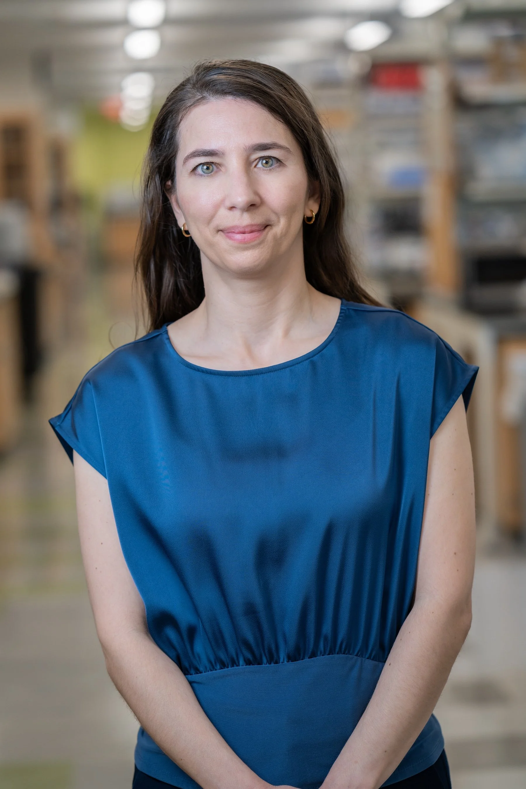 Portrait of a young woman with long brown hair, wearing a navy blue shirt, smiling outdoors with blurred greenery background.