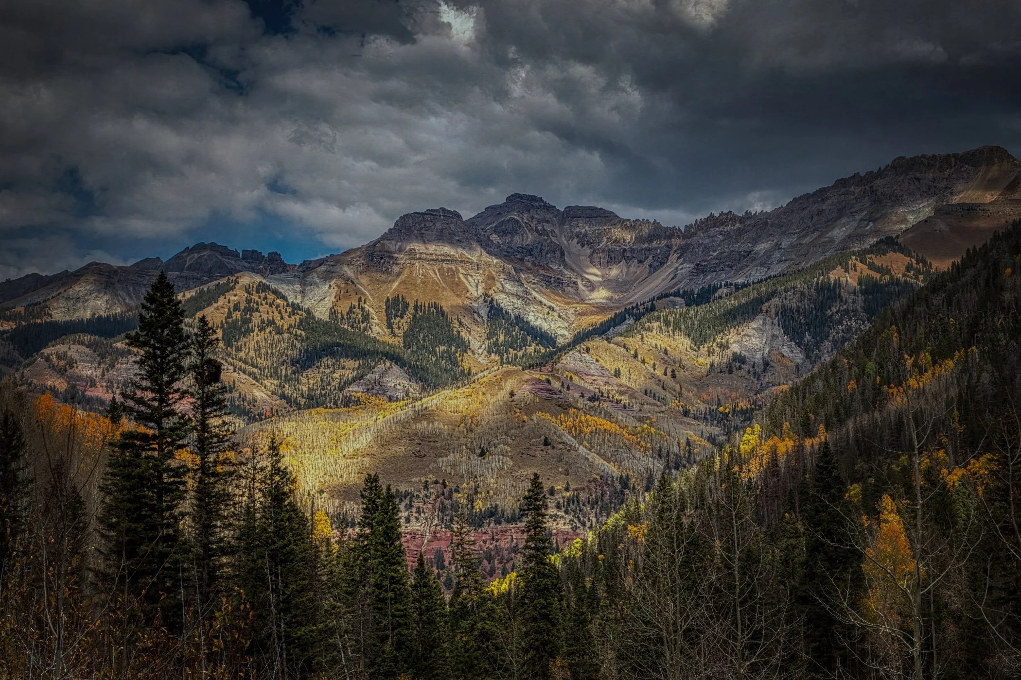 A mountain landscape showing rugged peaks under a dark, cloudy sky, with forested slopes and some autumn foliage in the foreground.