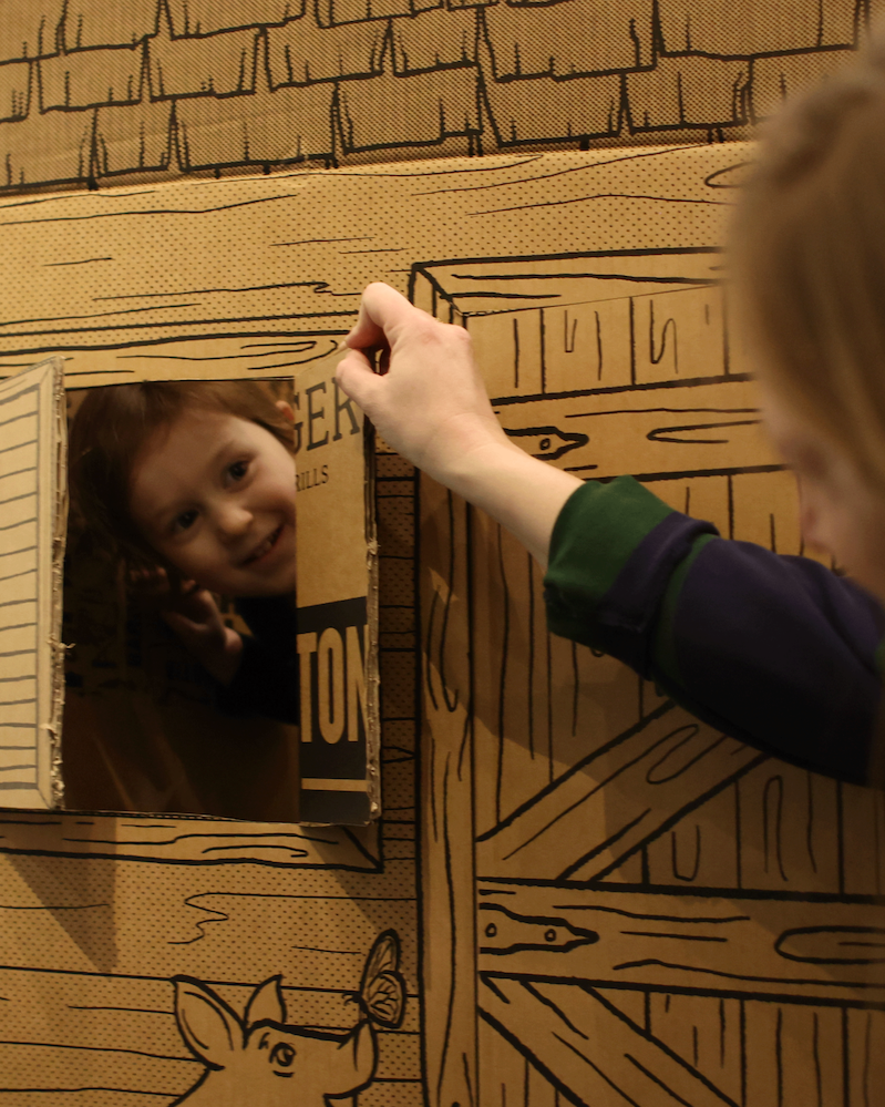 Child with a green and navy jacket looking into a small mirror on a cardboard wall decorated with barnyard illustrations.