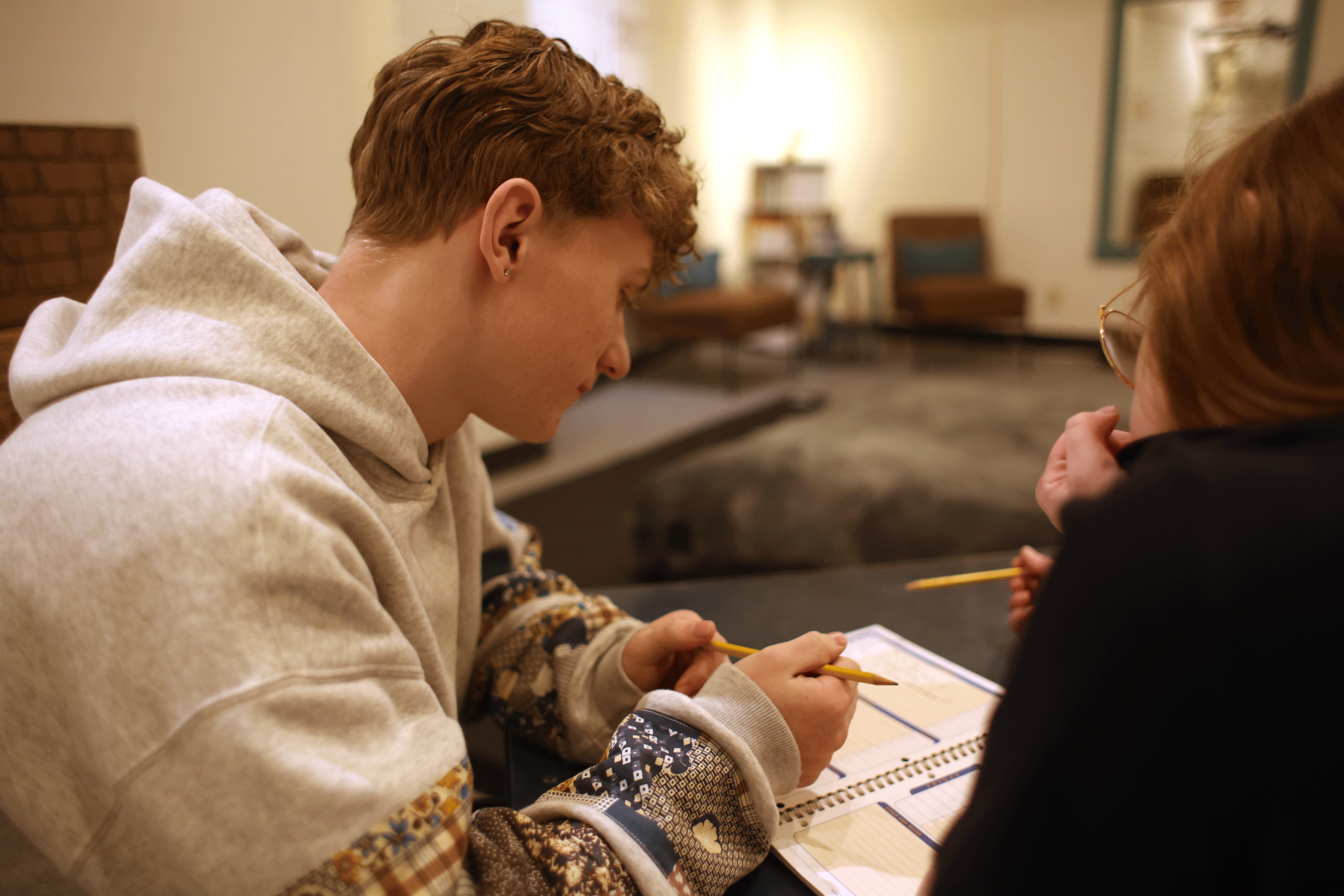 Two people sitting at a table, engaged in writing or drawing in notebooks, with a background of a furnished room.