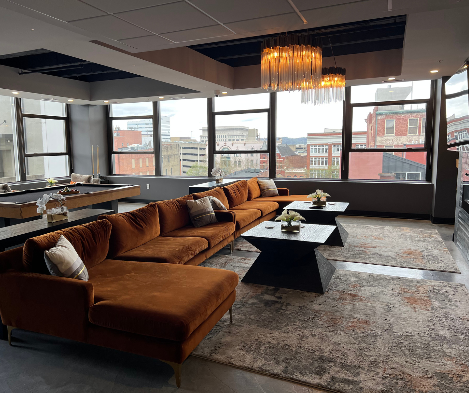 Modern high-rise lounge with large windows, a brown sectional sofa with pillows, black marble coffee tables with decorative flowers, a patterned area rug, and a statement chandelier.